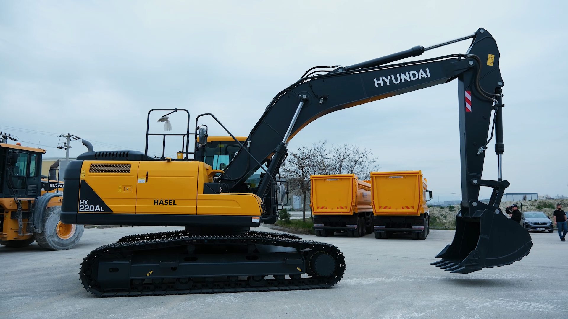 A large yellow and black Hyundai HX 220AL excavator with a bucket, parked outdoors with dump trucks.
