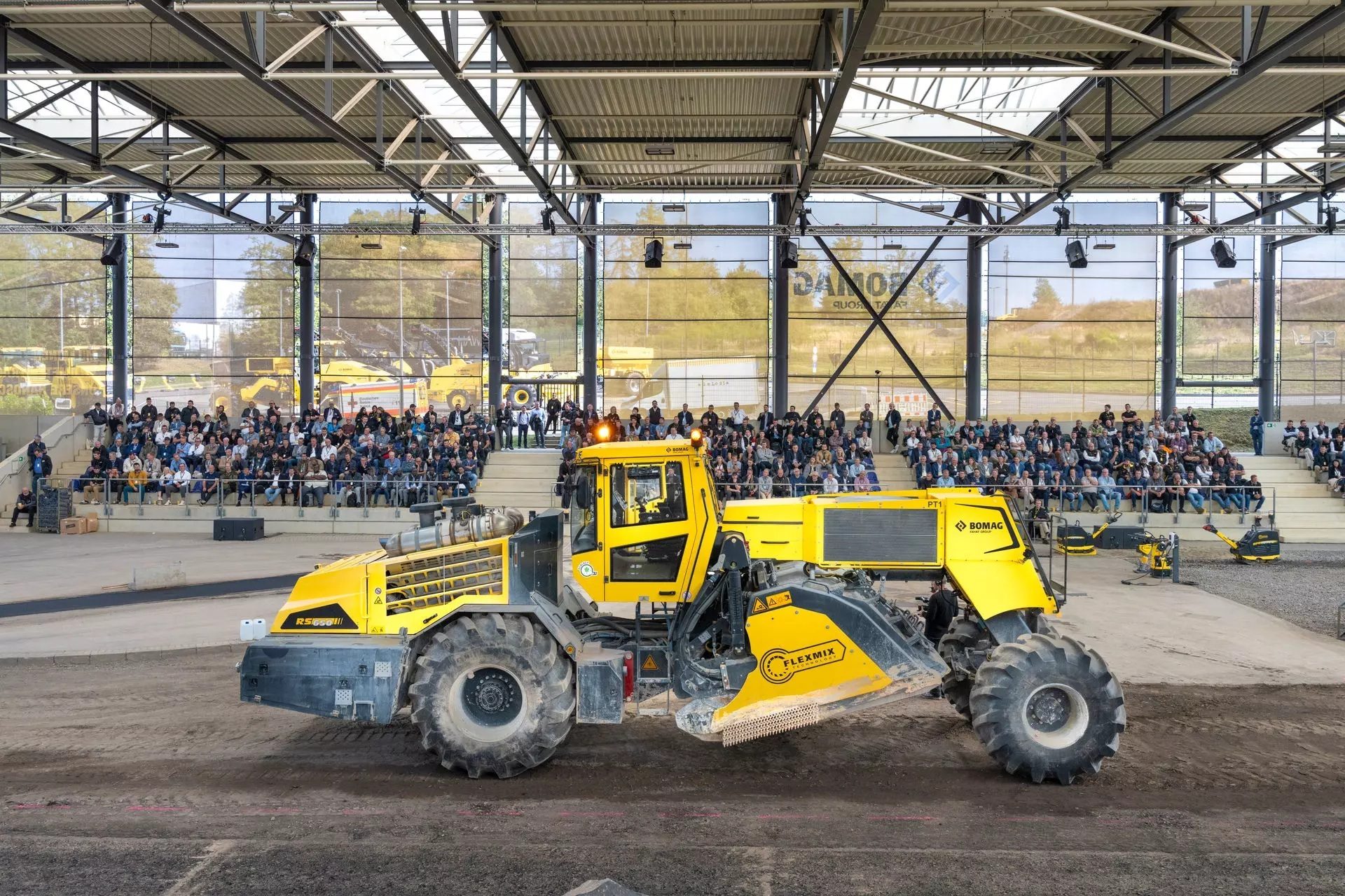 Large yellow road construction machine demonstrating for an audience in a spacious indoor arena.