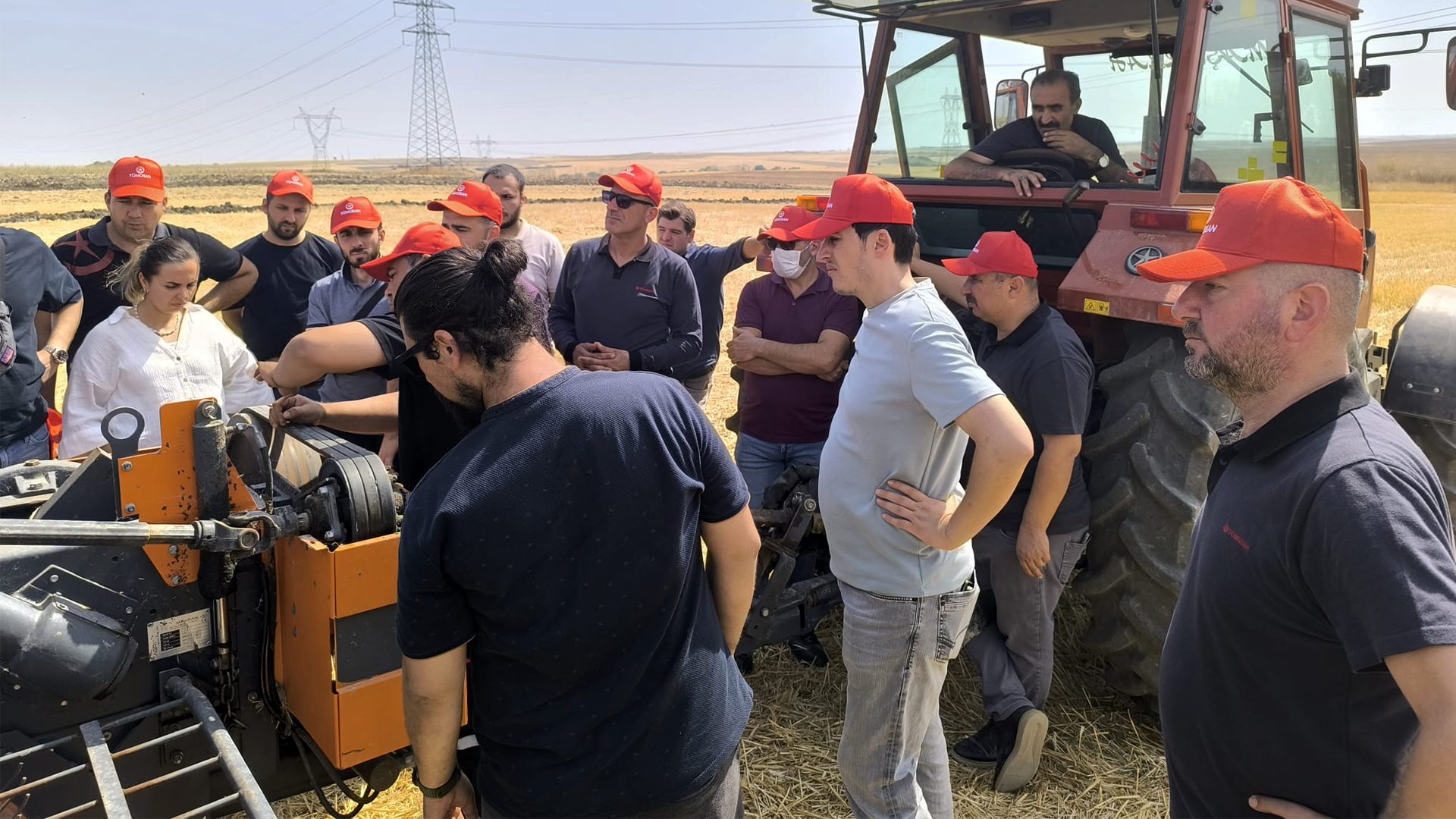 A group of people, many in red hats, inspecting farm machinery and a tractor in a field.