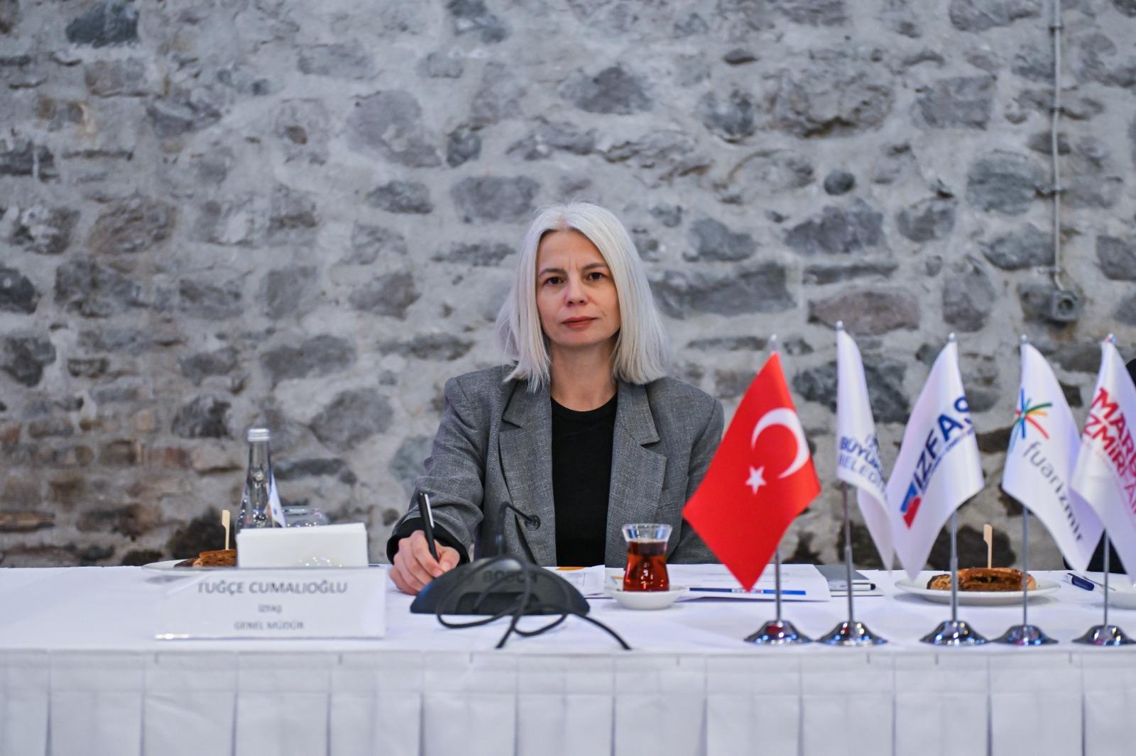 Woman, Tuğçe Cumalıoğlu, seated at a table with flags and a pen, against a stone wall.