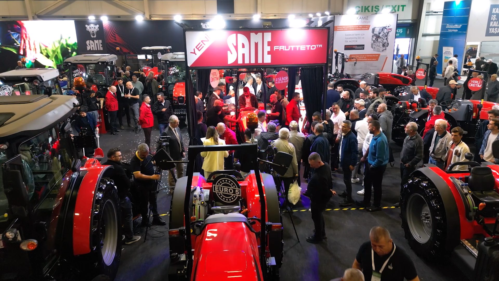 Busy indoor exhibition hall with many people gathered around red SAME brand tractors.