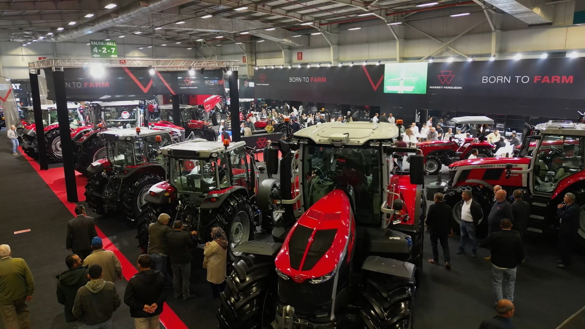 Indoor trade show with many red Massey Ferguson tractors and attendees, under "Born to Farm" banners.