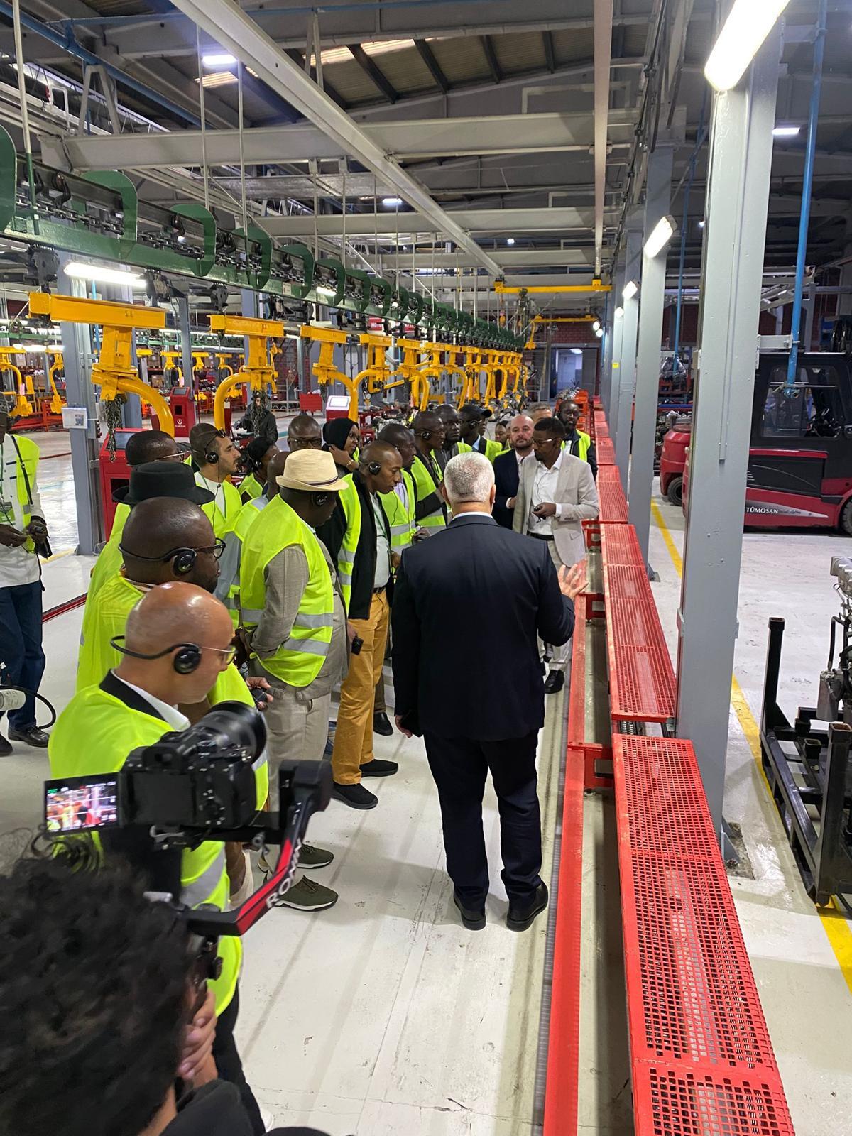A group of people in high-vis vests and headphones tour a factory with machinery, filmed by a cameraman.