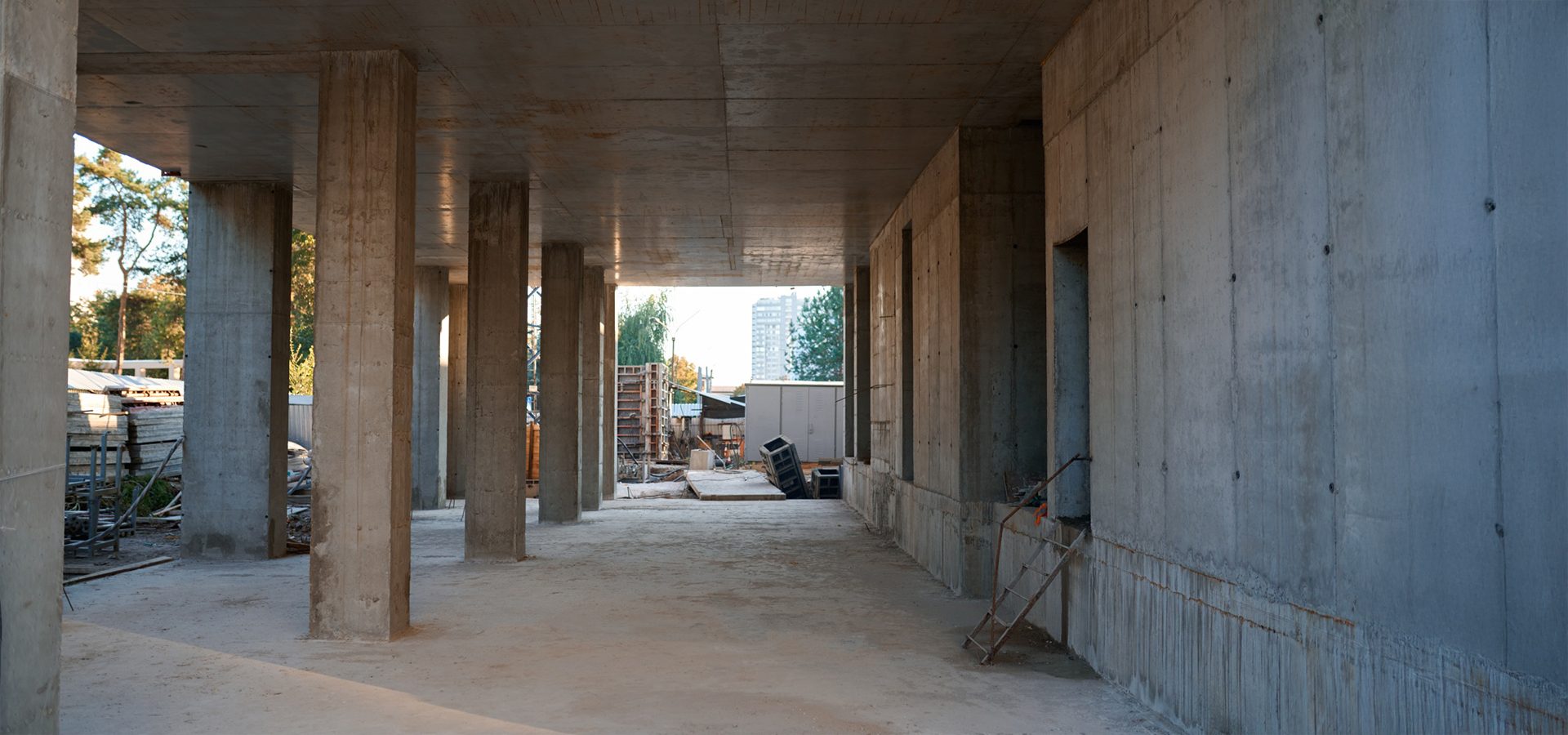 Interior view of an unfinished concrete building with pillars, walls, and a glimpse of an outdoor construction site.