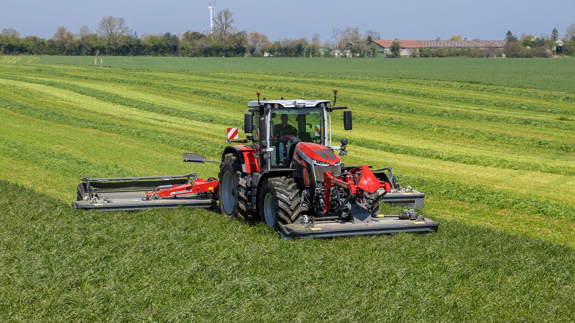 A red tractor with mowers cuts grass in a field, creating distinct stripes.