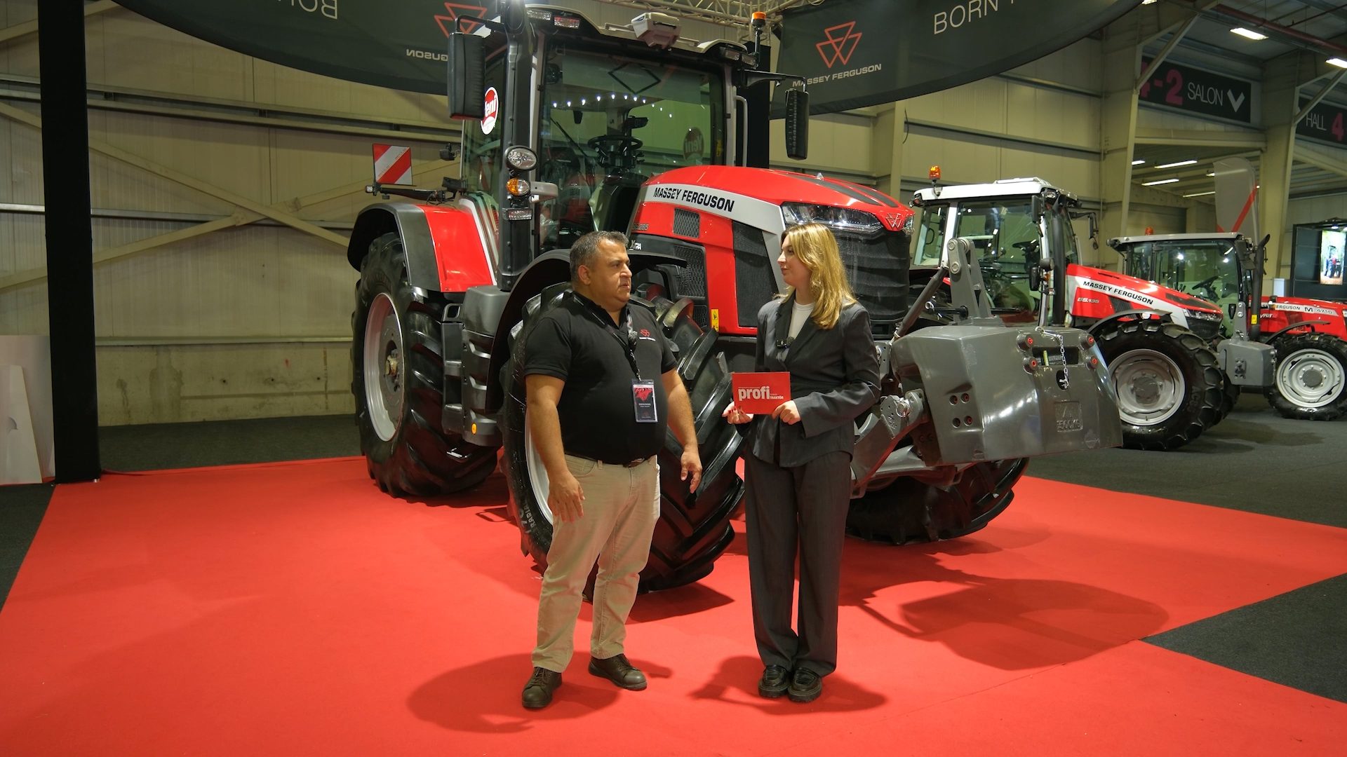 Two people stand before a red Massey Ferguson tractor at a trade show; the woman holds a "profi" sign.