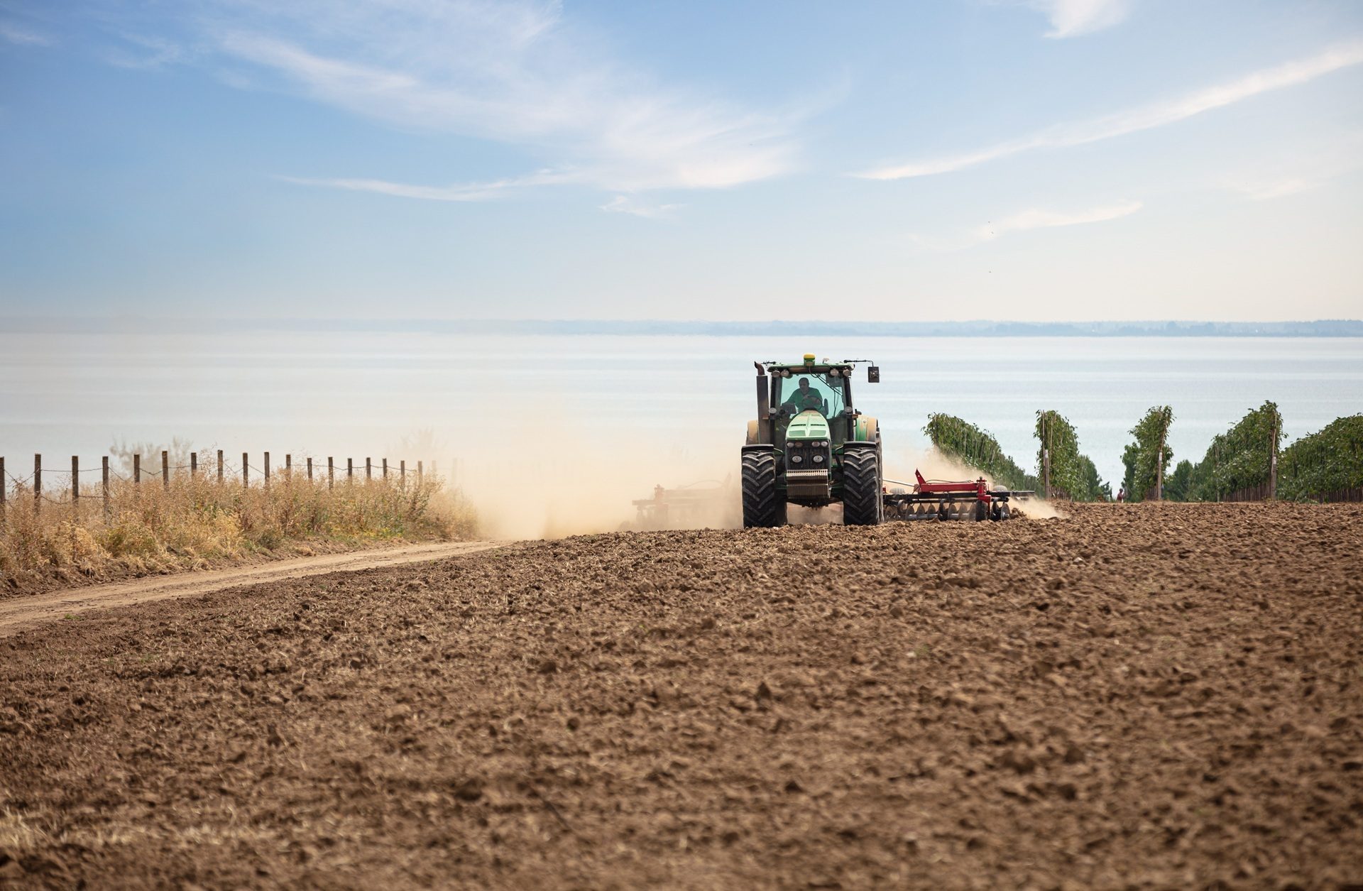 A green tractor plows a dusty field by a large lake, with a fence and vineyard rows.