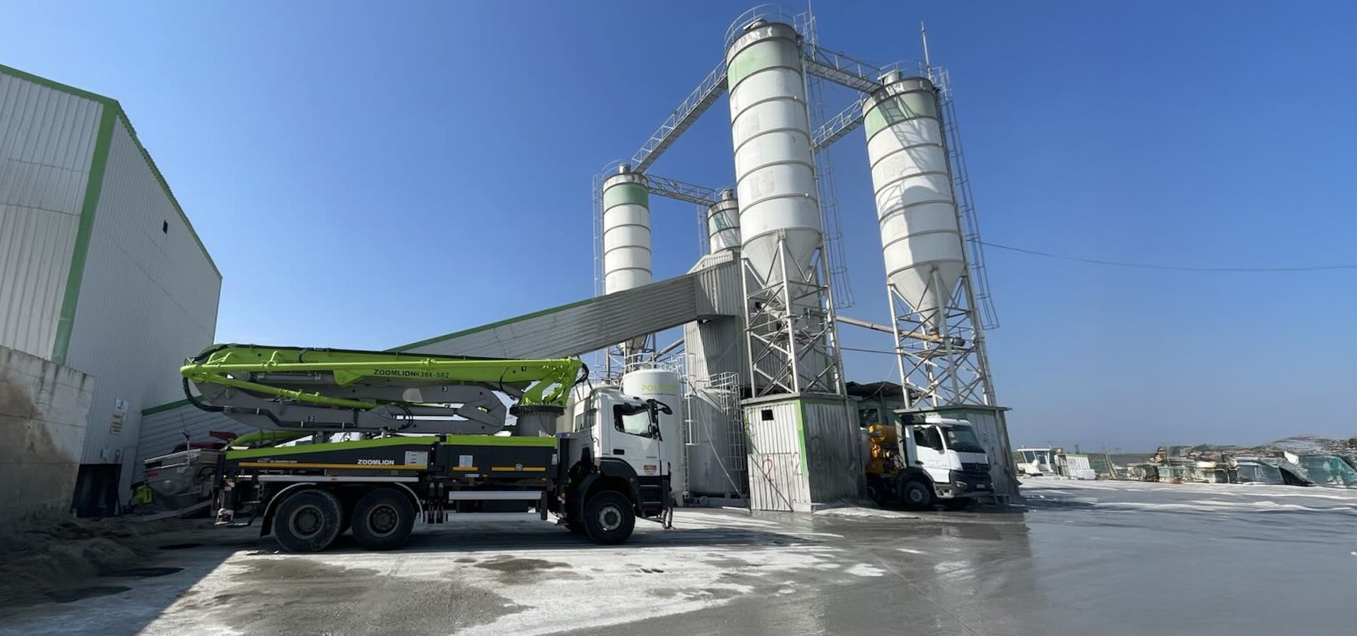 A ZOOMLION concrete pump truck is parked at a large concrete batching plant under a clear blue sky.