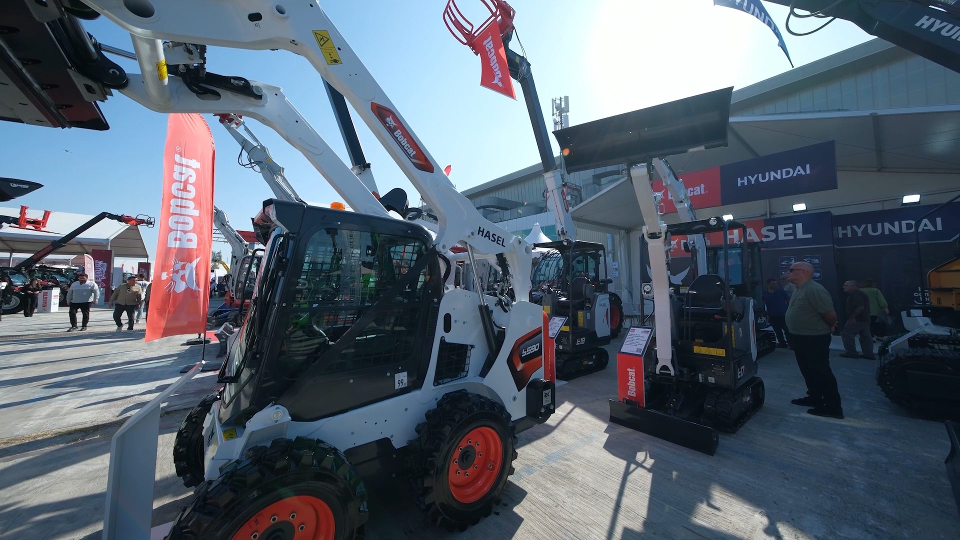 Outdoor exhibition of Bobcat skid-steer loaders and excavators with people, Hasel and Hyundai branding.