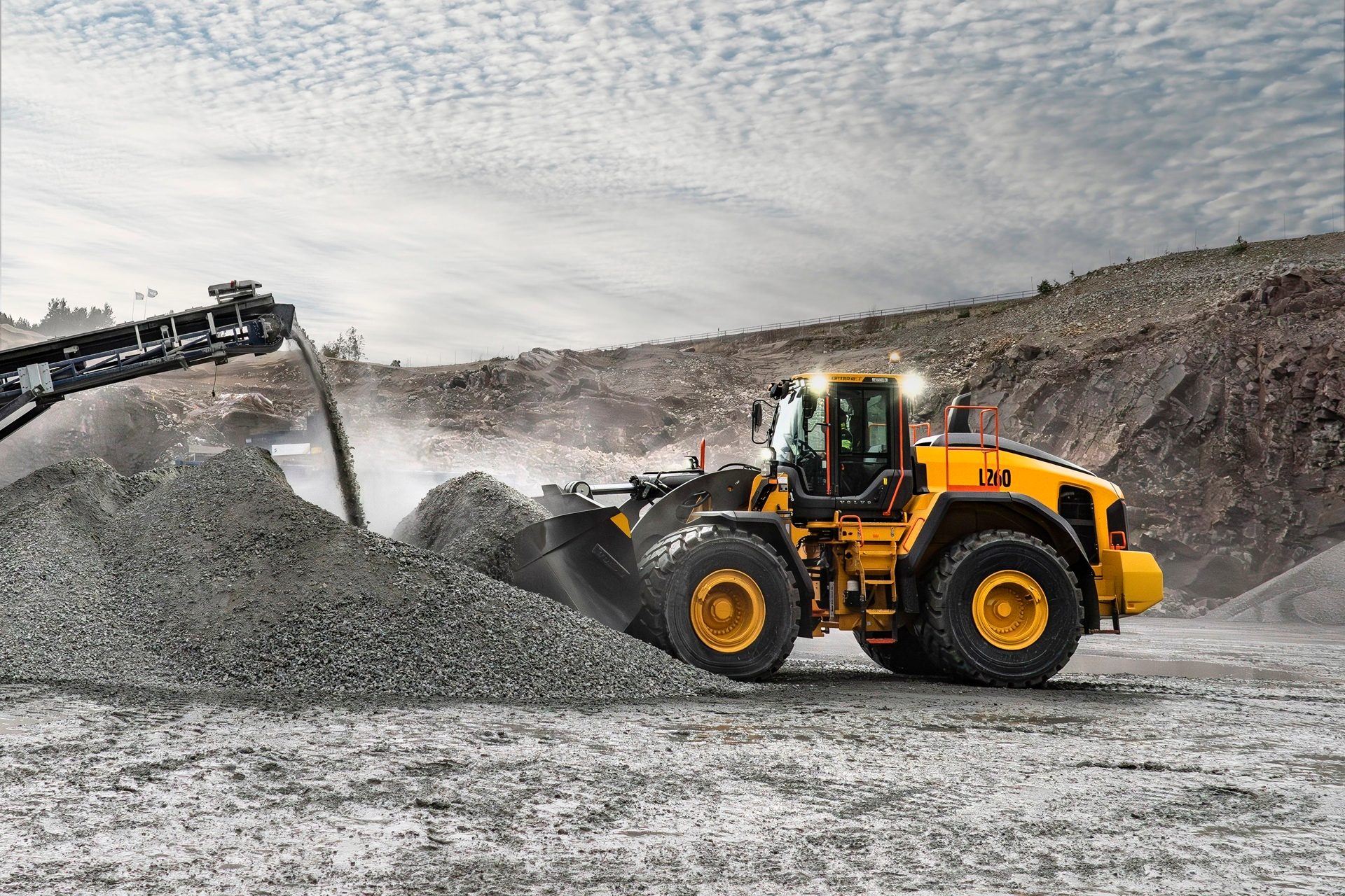 A large yellow wheel loader works alongside a conveyor belt, moving and crushing rock at a quarry.