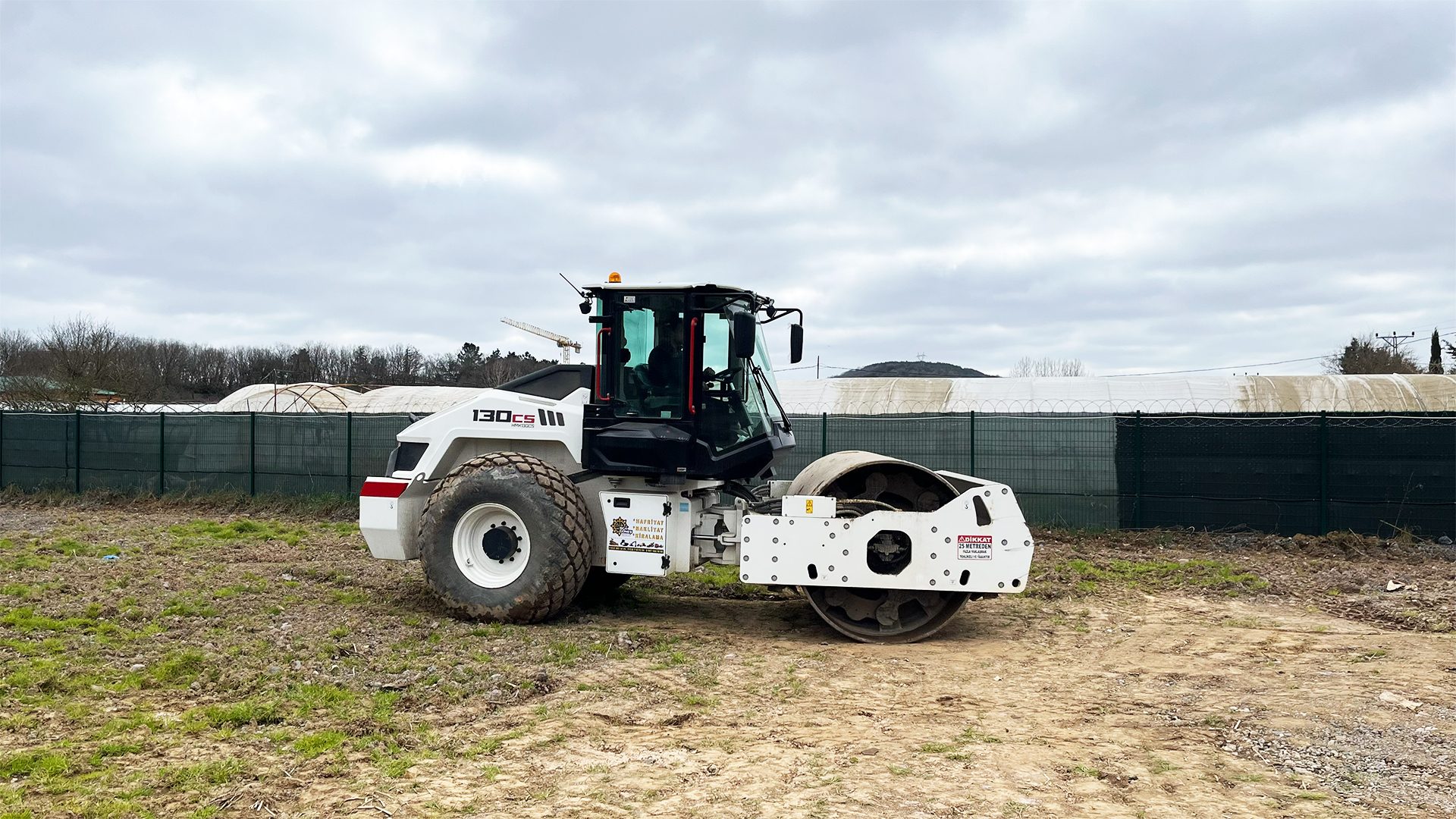 White 130cs road roller on dirt ground, with a green fence and cloudy sky in the background.