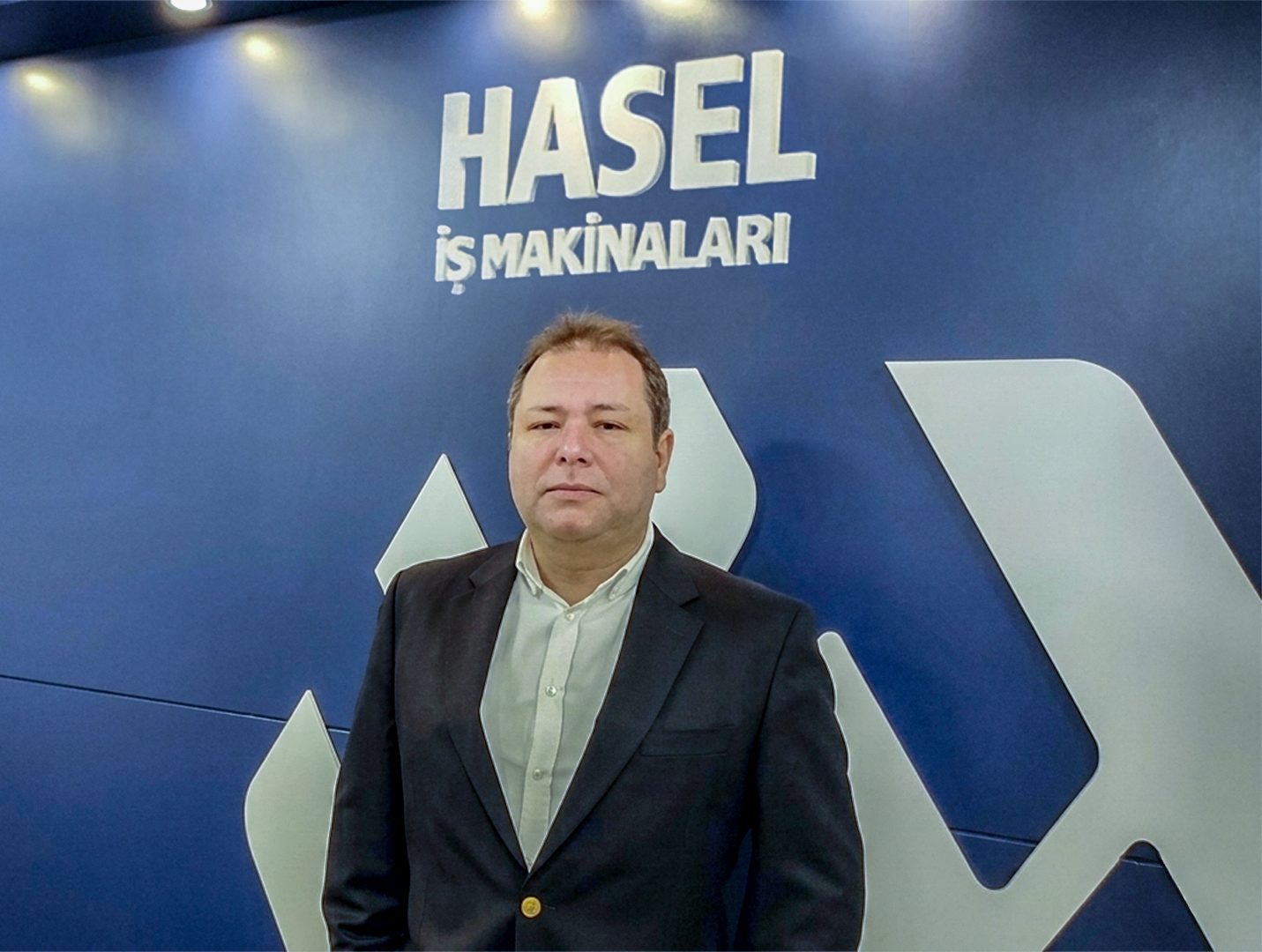 Man in a suit standing in front of a blue wall with the "HASEL İŞ MAKİNALARI" logo.