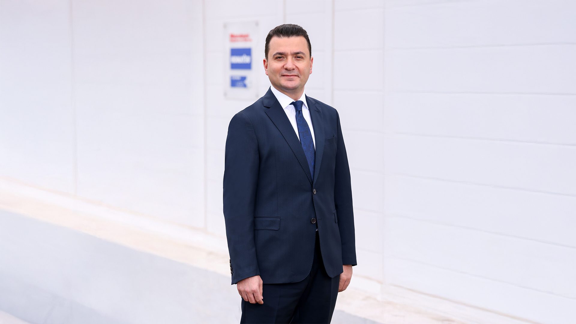 Smiling man in dark suit and blue tie stands against a white wall.
