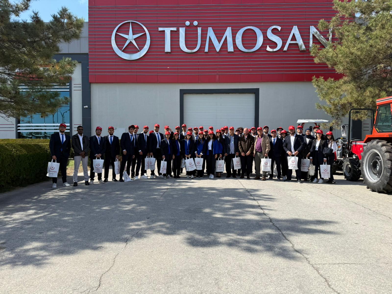 Group of people in red hats and suits stand in front of the TÜMOSAN building with tractors.