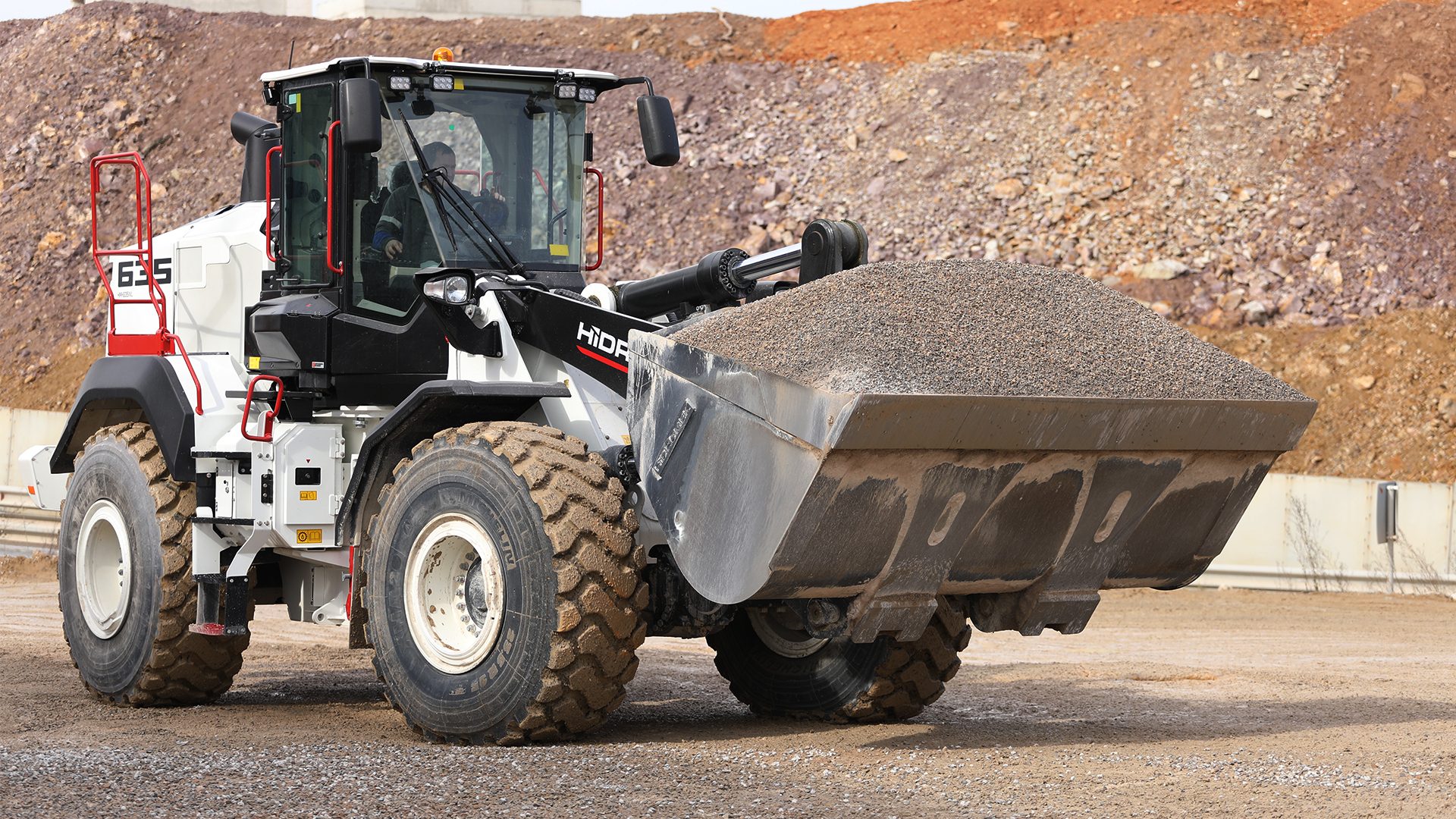 White front-end loader with a full bucket of gravel on a dirt road at a construction site.