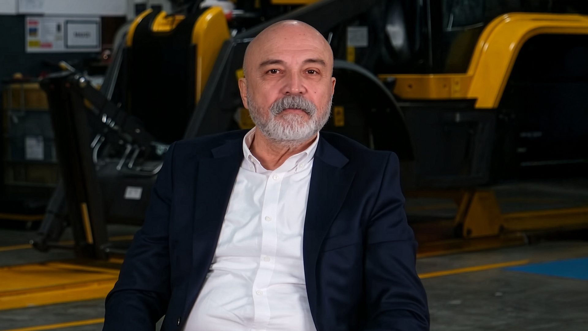 Bald, bearded man in a suit sits in an industrial setting with yellow and black machinery.