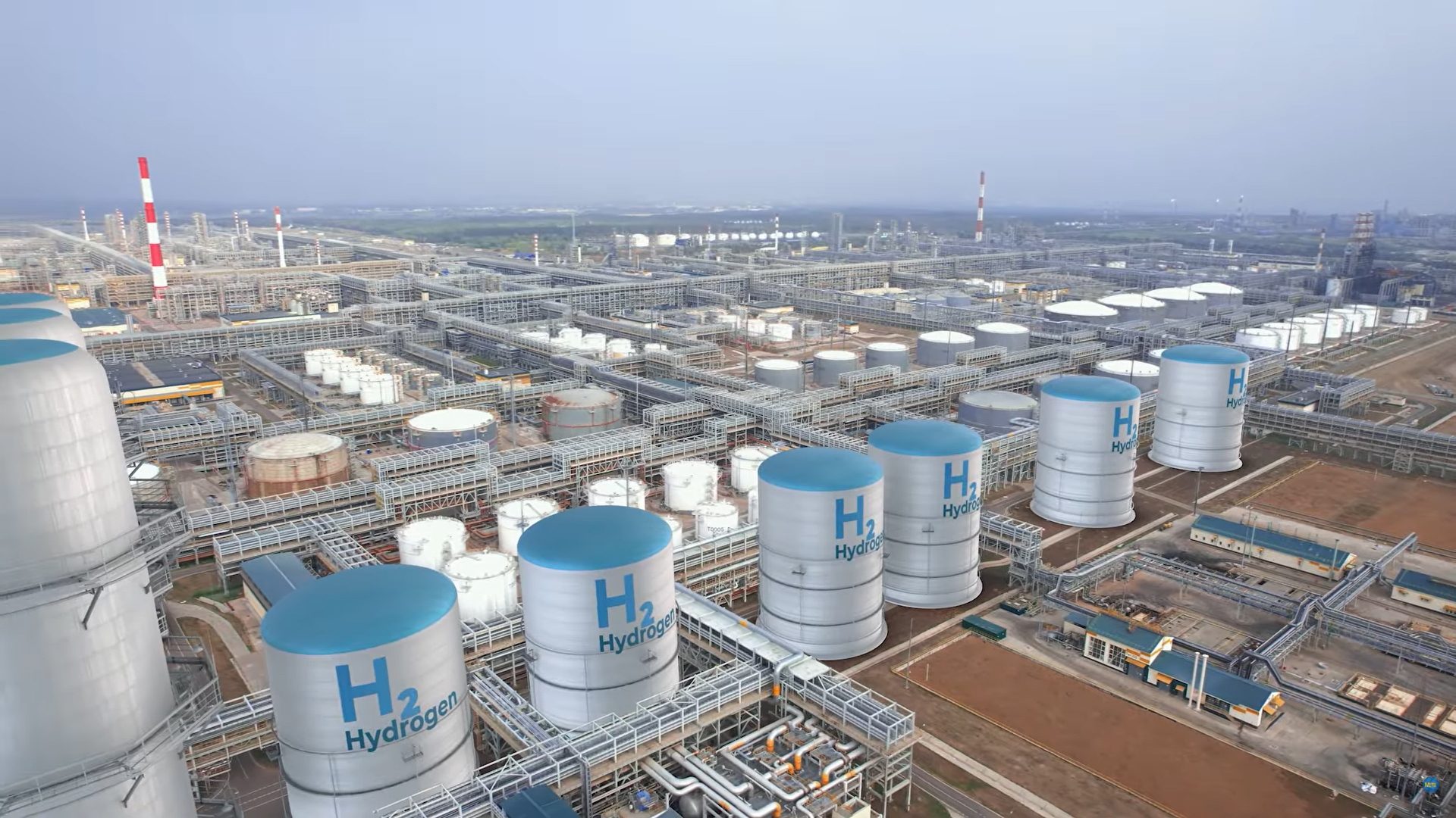 Aerial view of a large industrial complex with many cylindrical storage tanks labeled "H2 Hydrogen" and pipelines.