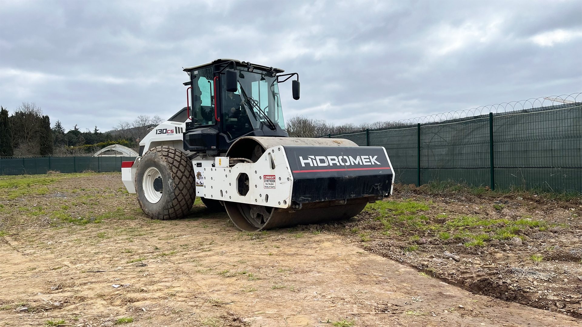 A white and black Hidromek 130cs soil compactor parked on a dirt and grassy construction site under a cloudy sky.