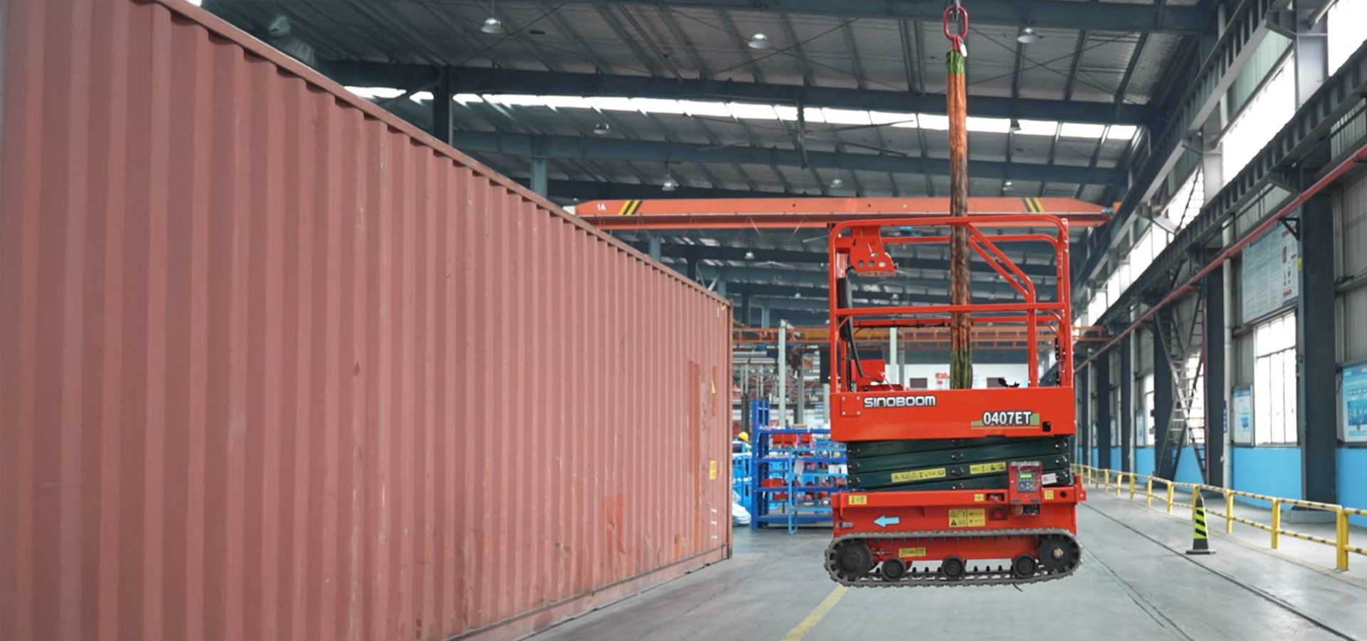 An orange Sinoboom 0407ET scissor lift is suspended by a crane next to a red shipping container in a factory.