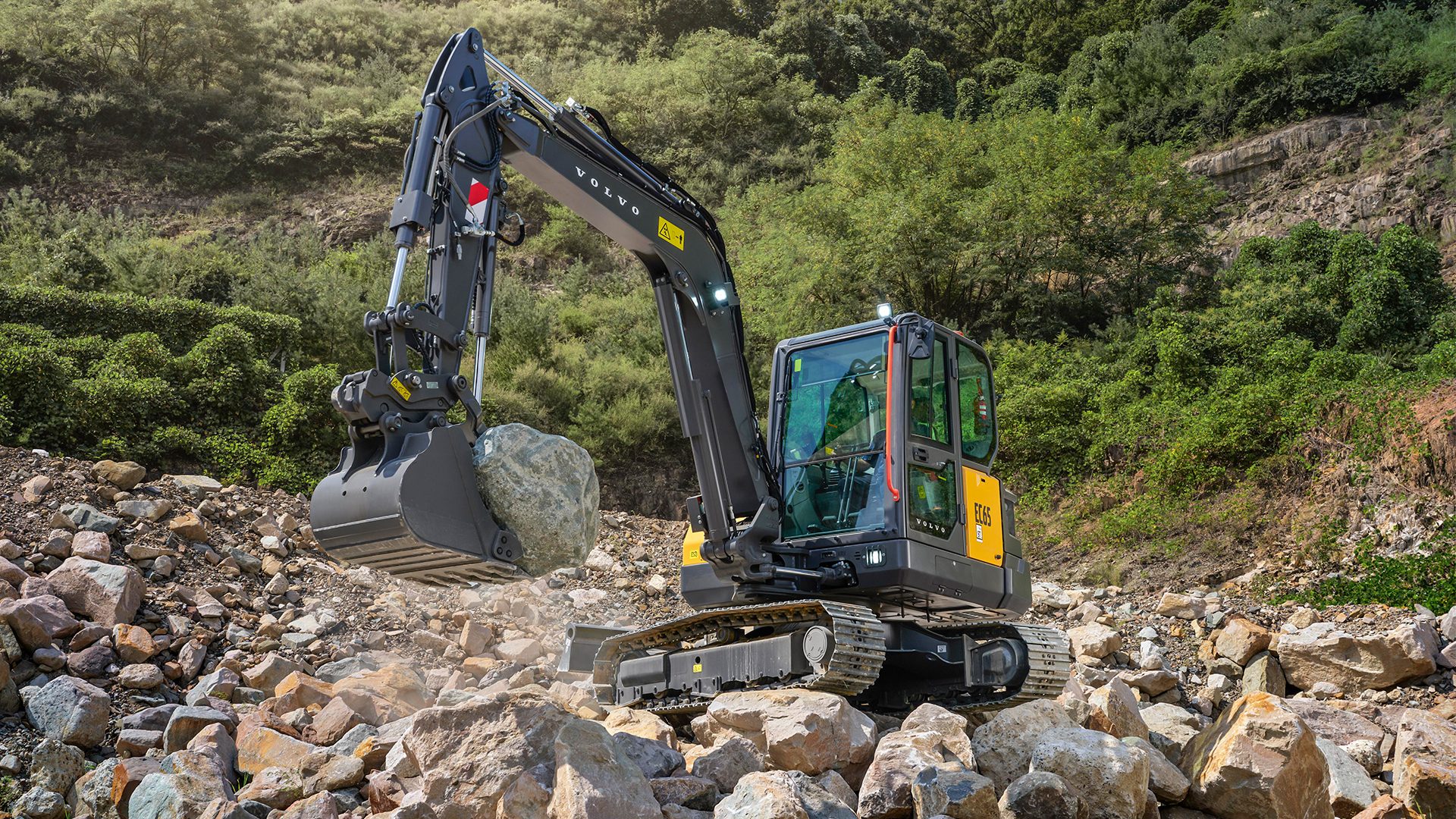 Volvo EC65 excavator moving a large rock on a construction site with rocky terrain and green hills.