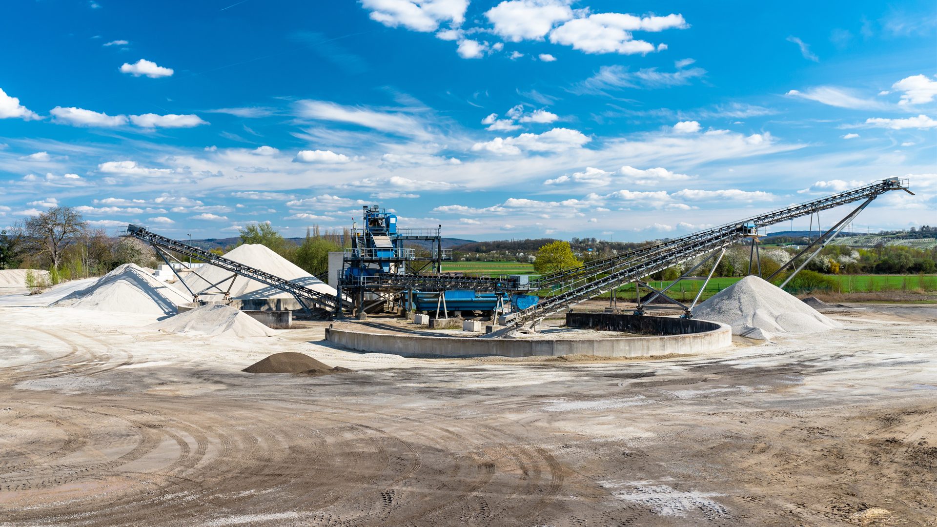 Industrial quarry scene: blue processing plant, long conveyor belts, sand/gravel piles, blue sky.