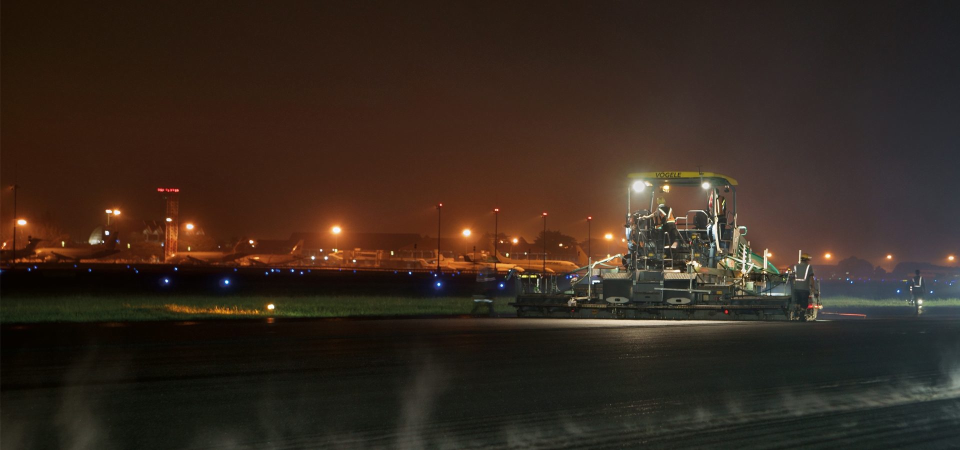 Night work: Large paver and crew on an airport runway under bright lights.