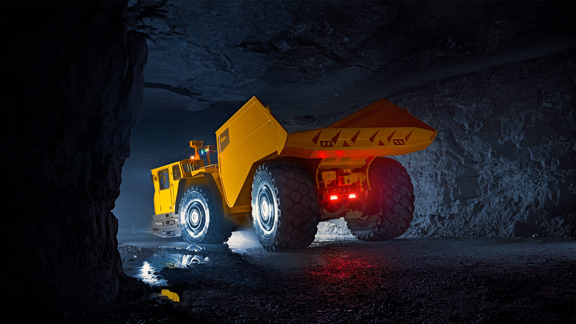 Large yellow mining dump truck with illuminated wheels and red taillights in a dark underground mine.