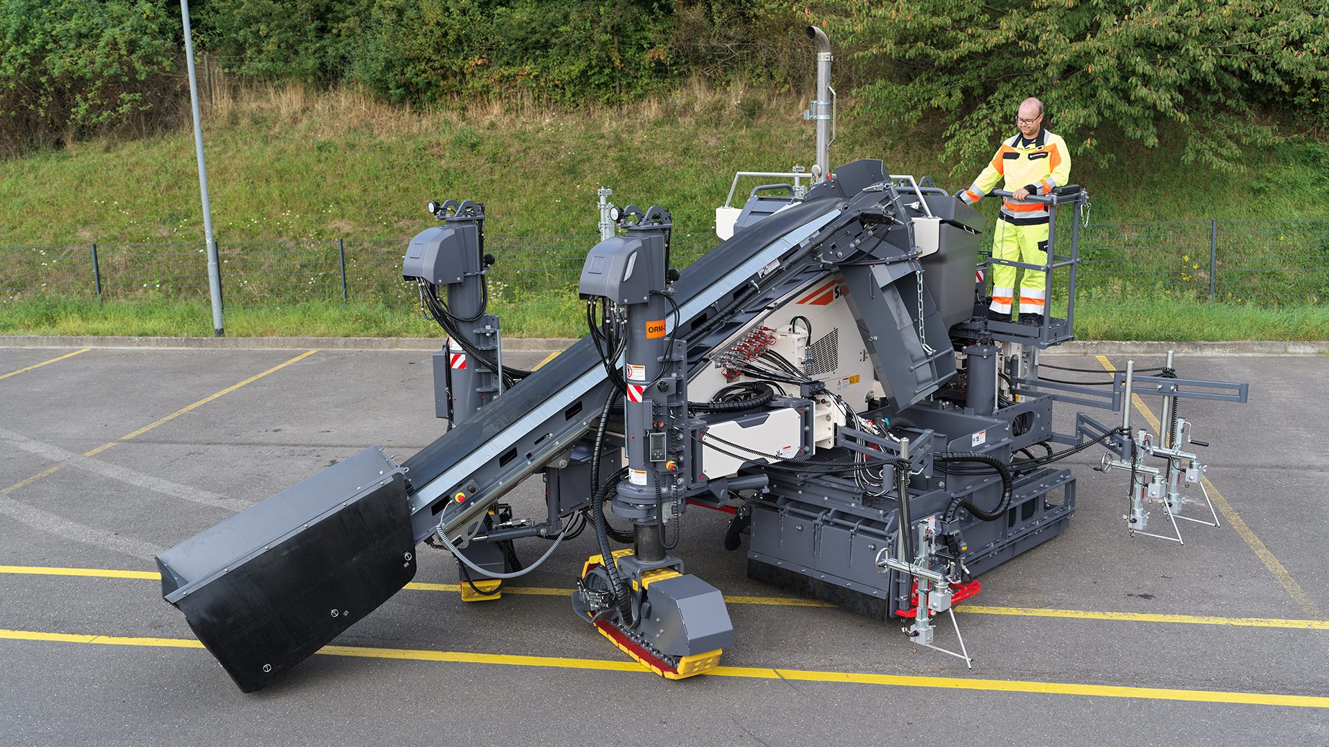A man in high-vis clothing on a large gray asphalt milling machine on a paved lot.