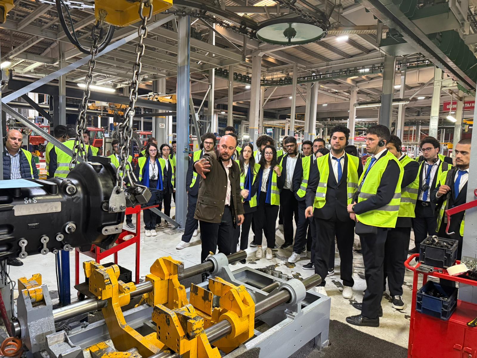 Students in safety vests on a factory tour, with a guide explaining machinery.