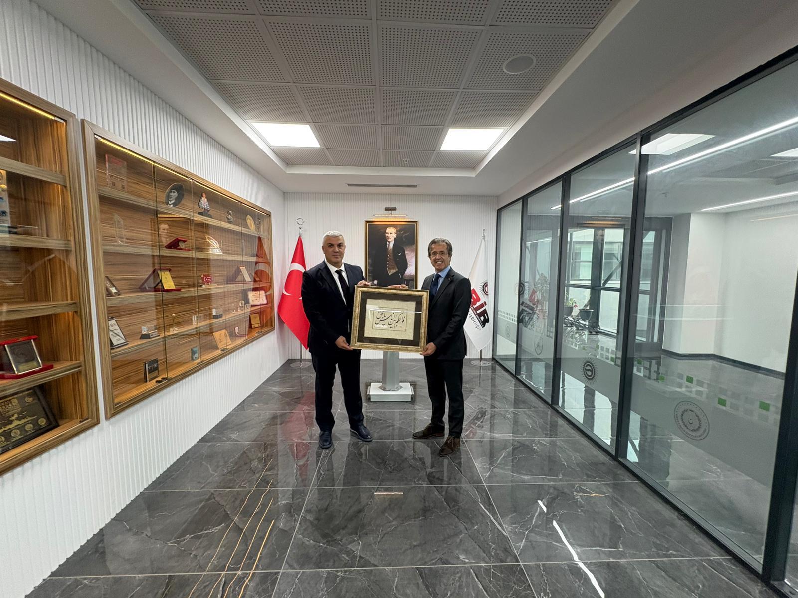 Two men in suits hold a framed calligraphic artwork in an office with Atatürk's portrait and Turkish flags.