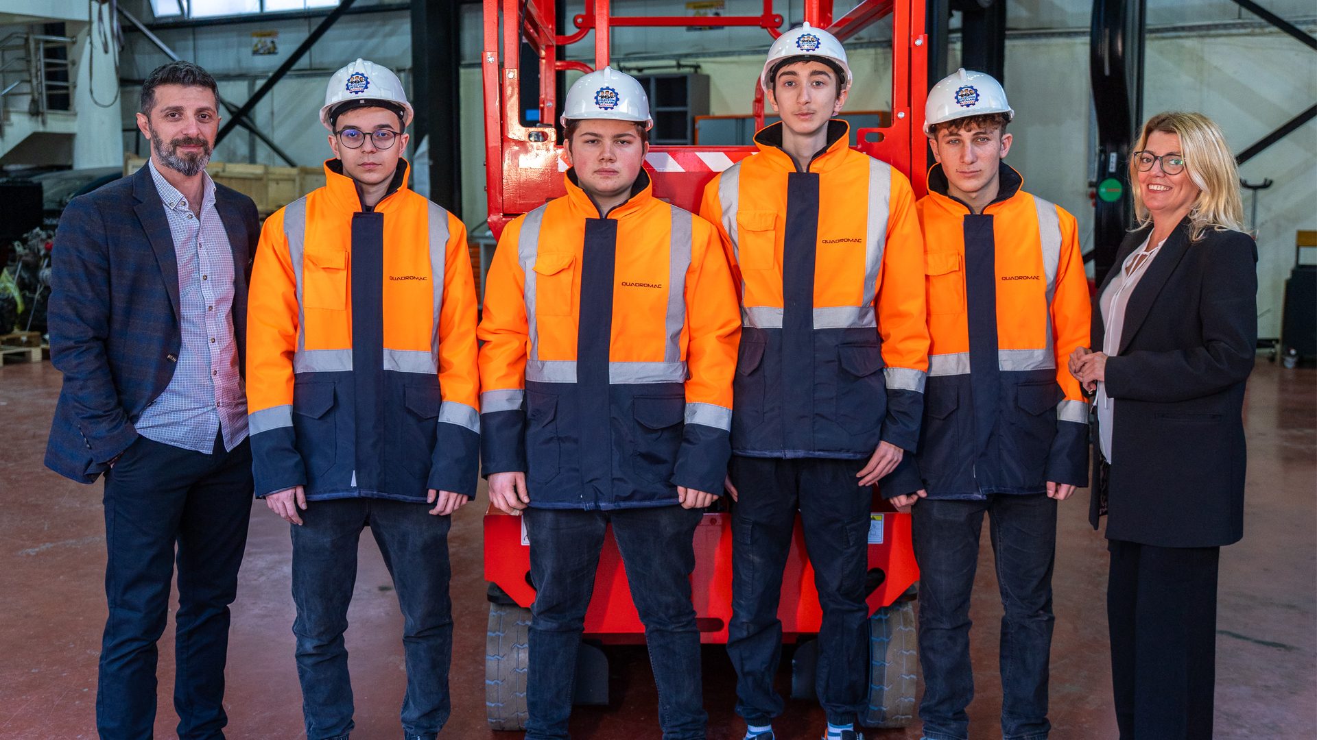 Six people, including four young men in safety gear, standing in front of industrial machinery.