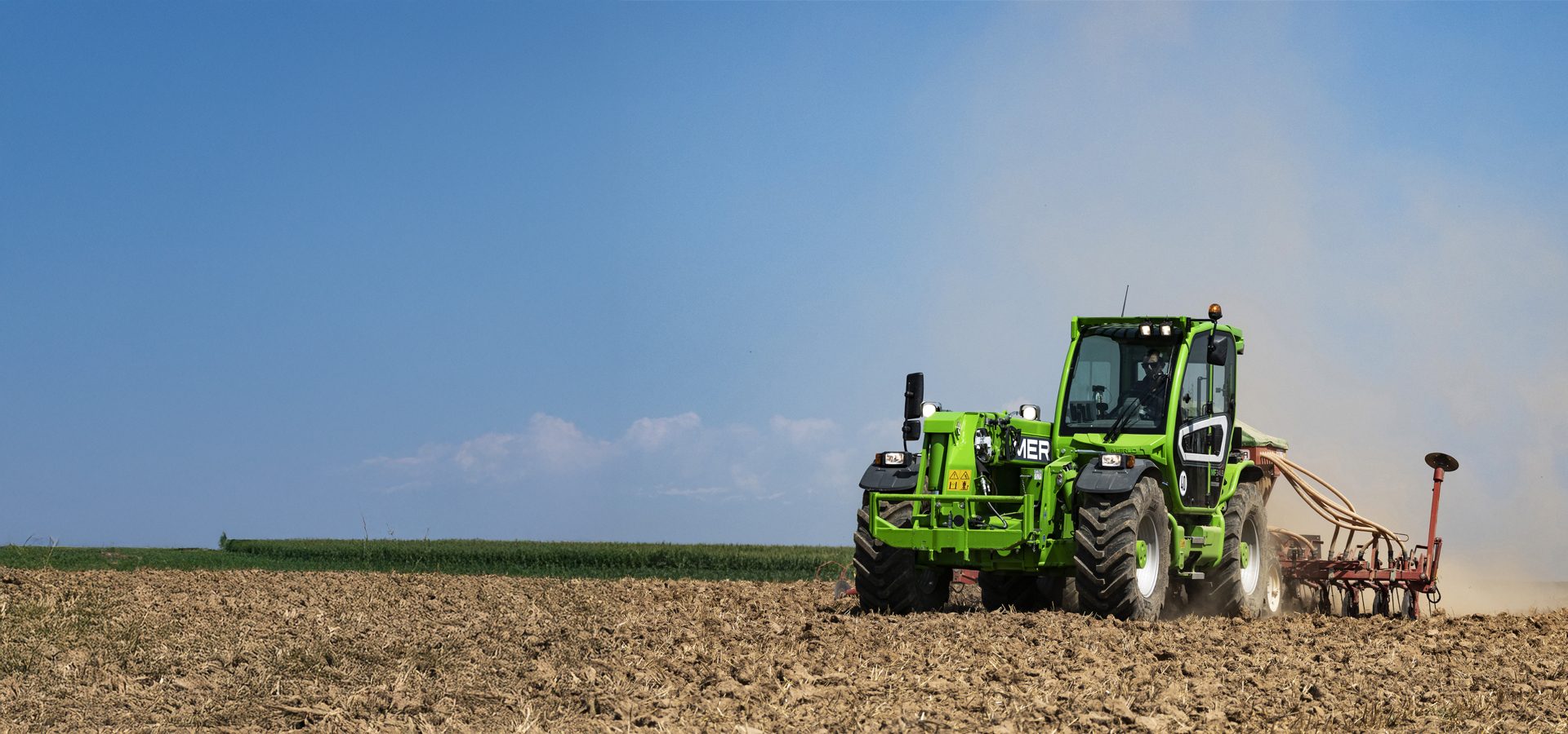Bright green telehandler seeding a brown field, dust rising under a blue sky.