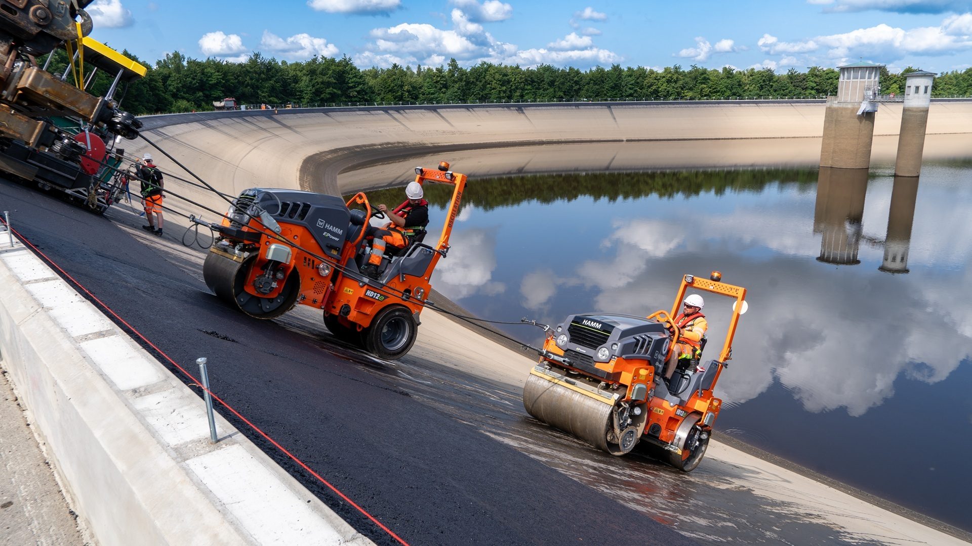 Construction workers pave a steep asphalt slope beside a reservoir with two orange rollers.