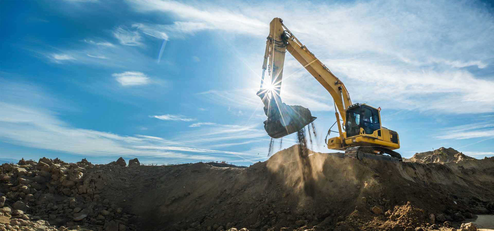 Yellow excavator dumping dirt under a bright sun and blue sky.