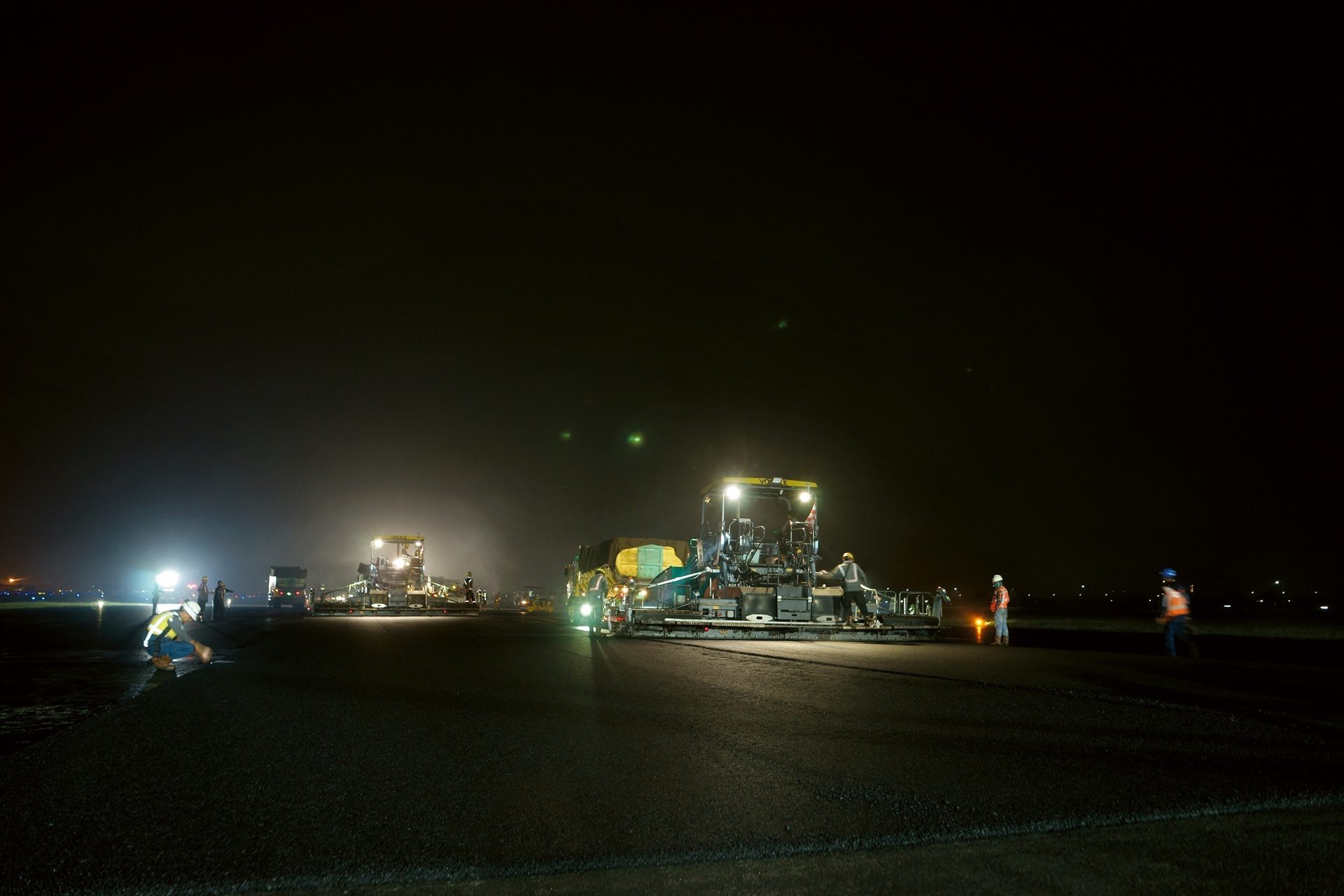 Nighttime road paving with large machines and workers, brightly lit.