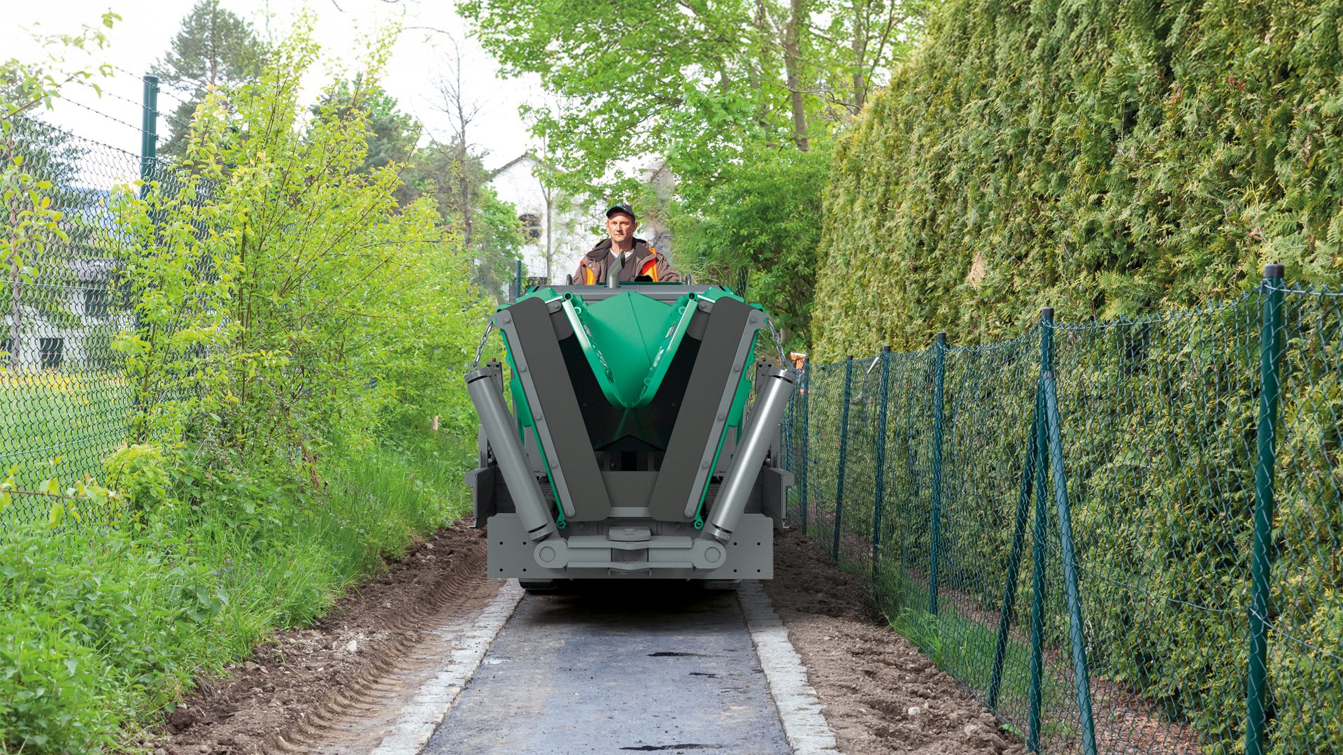 Man paving a narrow path with a green machine, flanked by fences and greenery.