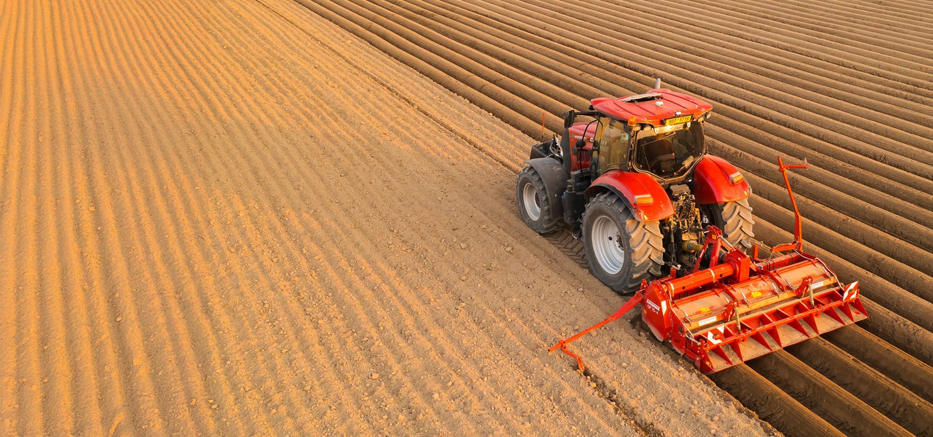 Red tractor tilling a field, creating parallel soil rows.