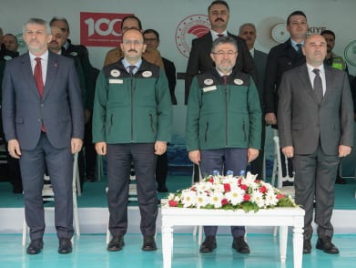 Group of men in suits and green jackets stand formally with a banner and floral arrangement.