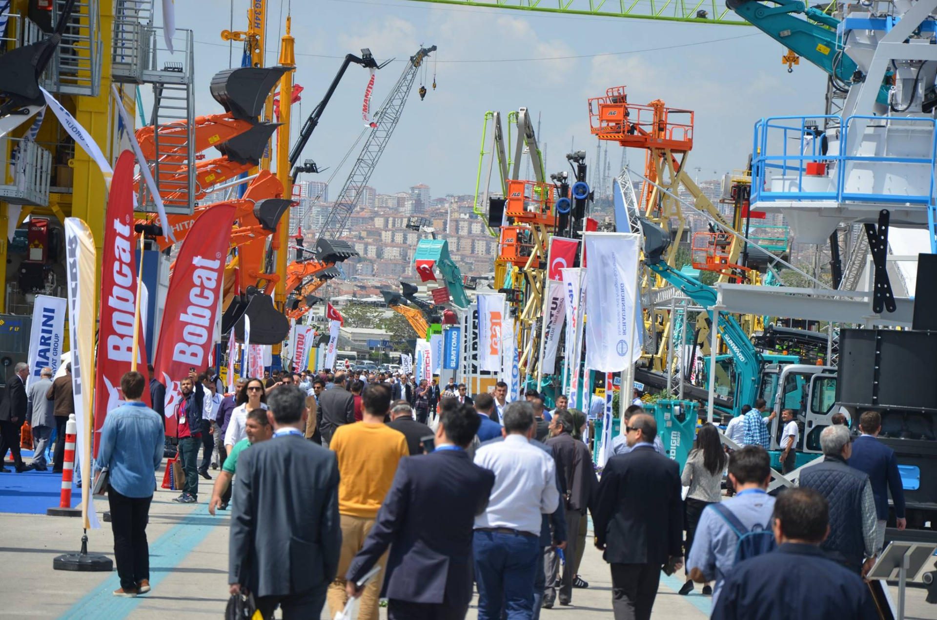 Outdoor trade show with people viewing heavy construction equipment, cranes, and brand flags.