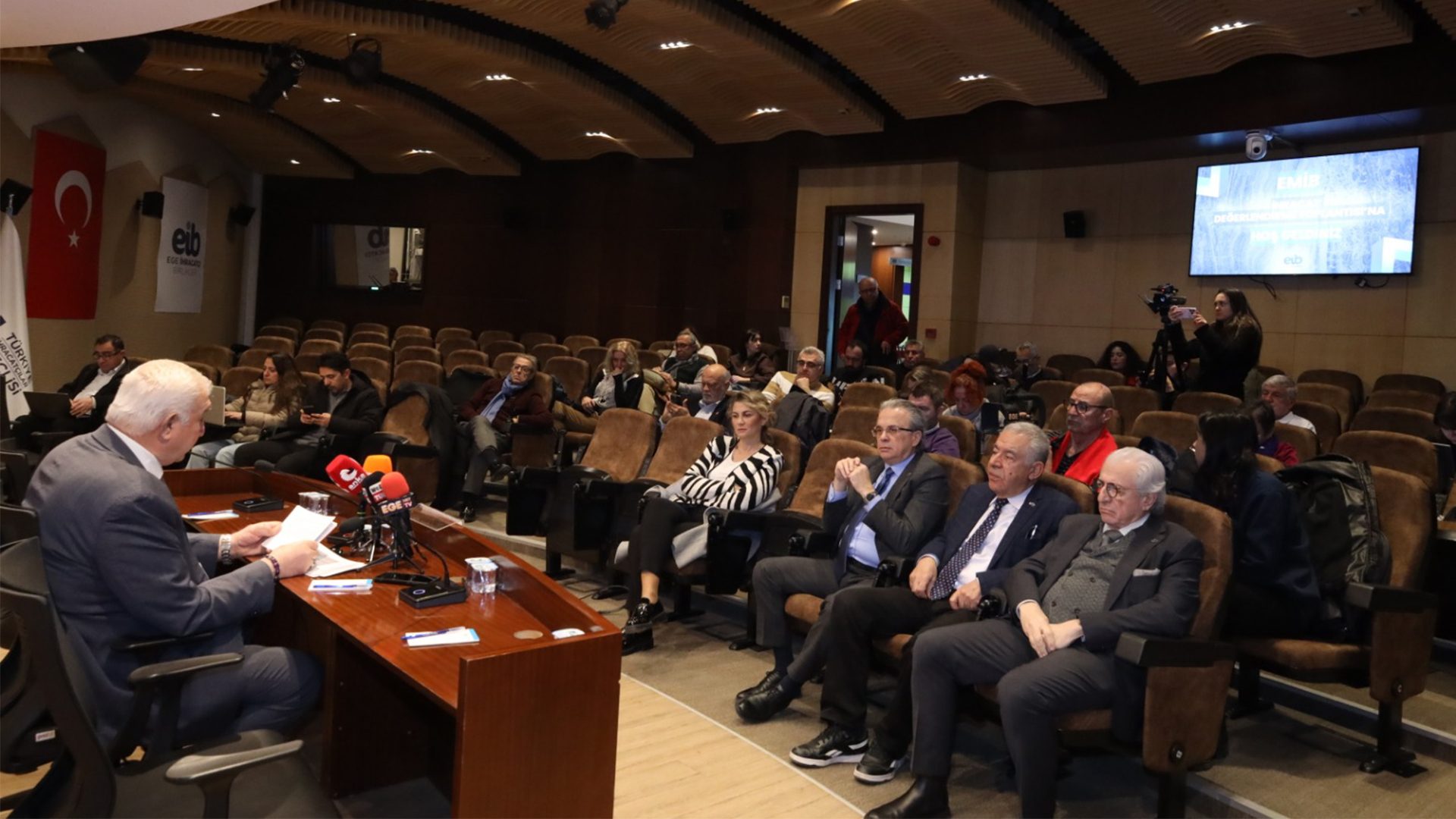 A speaker addresses an audience in a conference room with a large screen and a camerawoman present.