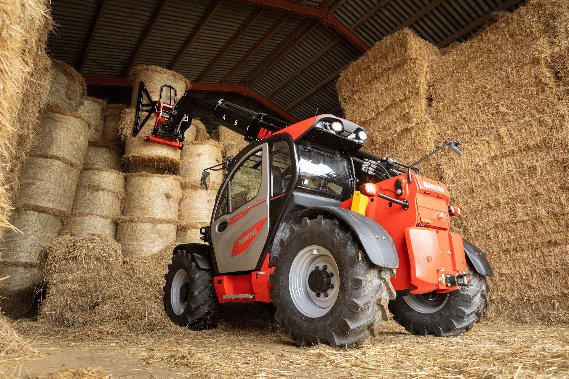 A red and grey Manitou telehandler moving hay bales inside a barn with high stacks of hay.