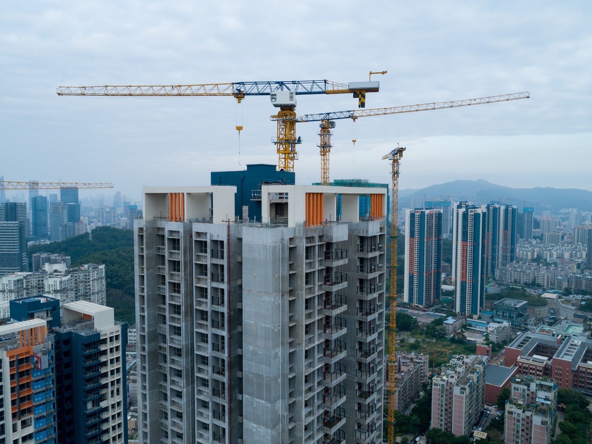 An aerial view of a tall, unfinished concrete building under construction with multiple tower cranes against a city skyline.