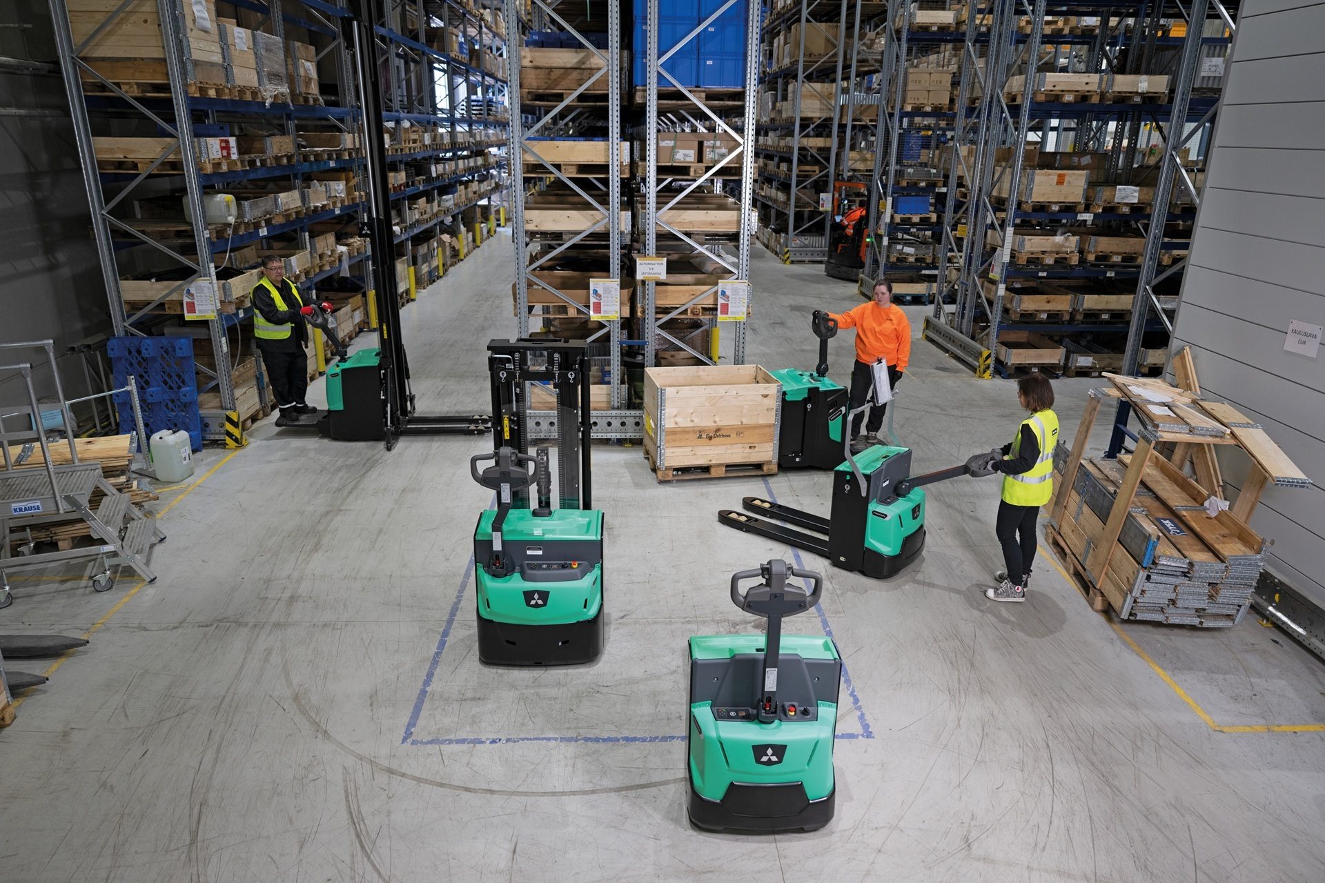 Warehouse workers operate pallet jacks and forklifts among storage racks, moving goods.