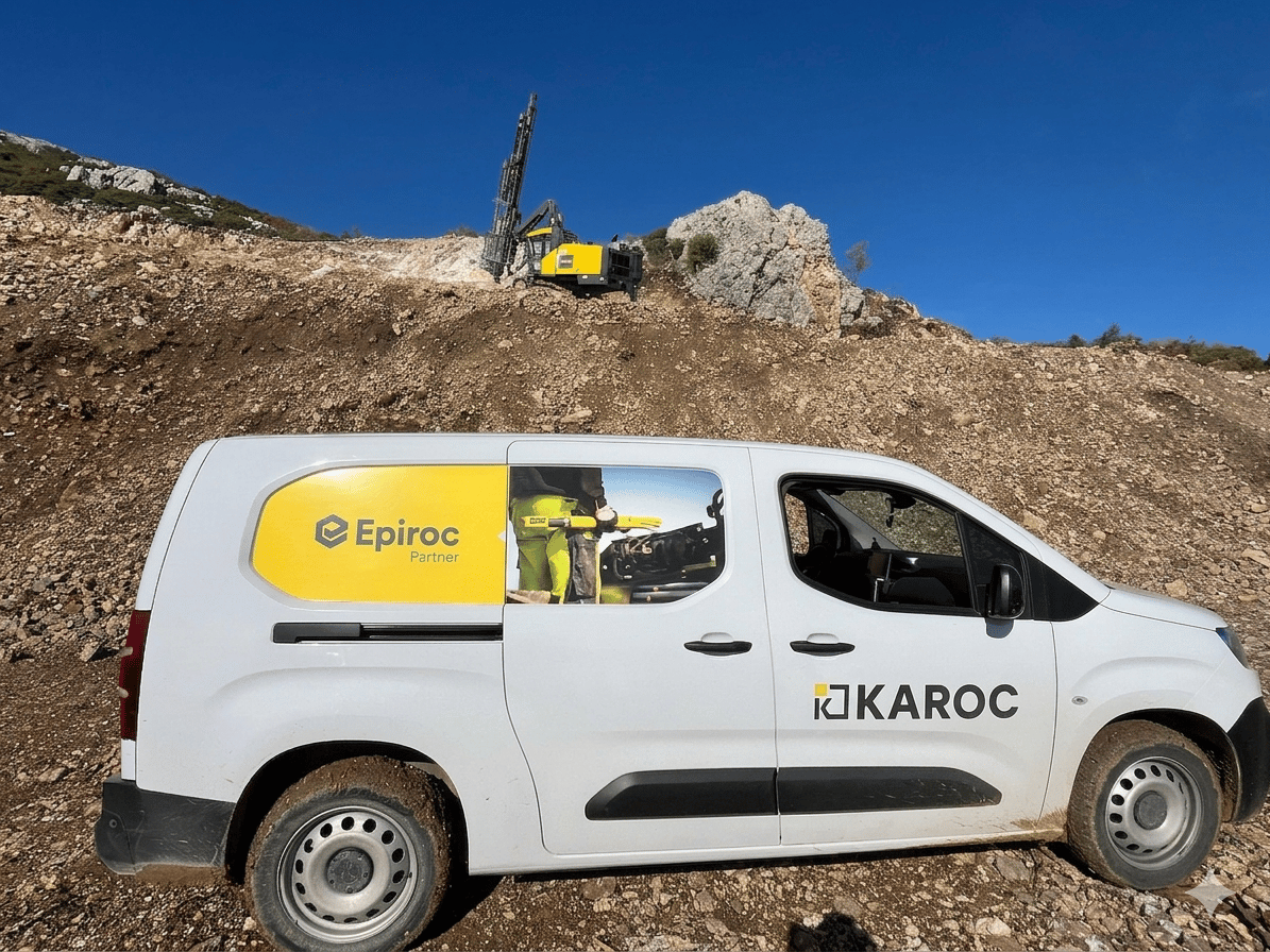 White van with Epiroc and KAROC logos and a yellow drilling rig on a rocky terrain under blue sky.