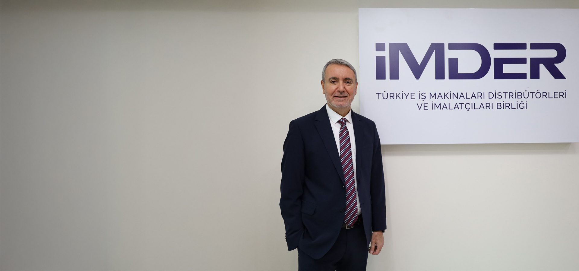 A smiling man in a suit stands in front of a white wall with the "IMDER" logo and Turkish text.
