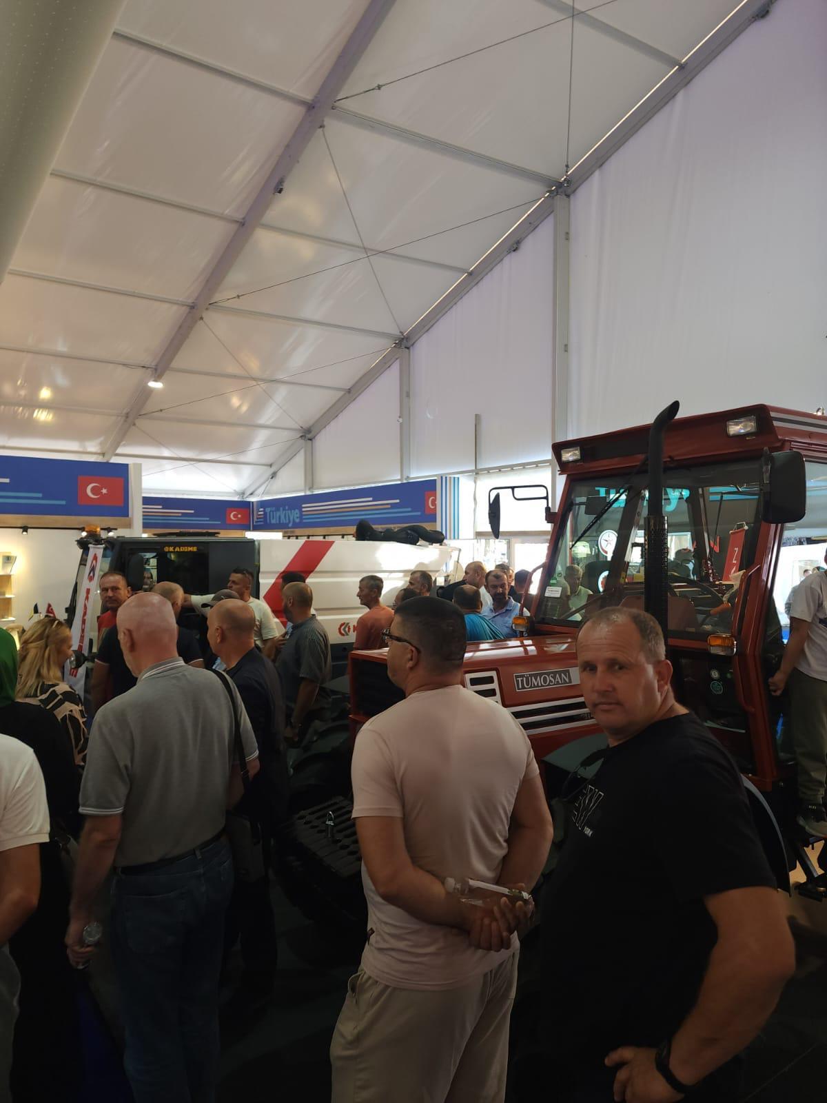 Crowd of men viewing a red TUMOSAN tractor and other farm equipment in an exhibition tent.