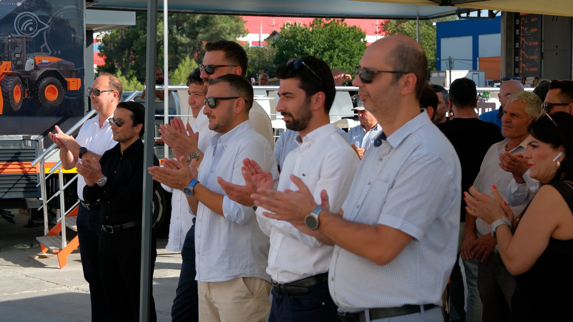 A diverse group of people clapping at an outdoor event, with heavy machinery in the background.
