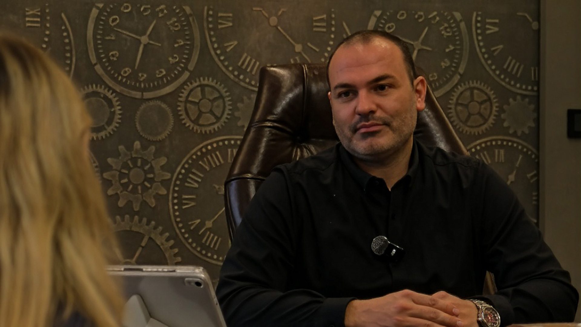 A man in a black shirt sits in a leather chair, facing a woman, against a steampunk-themed wall.