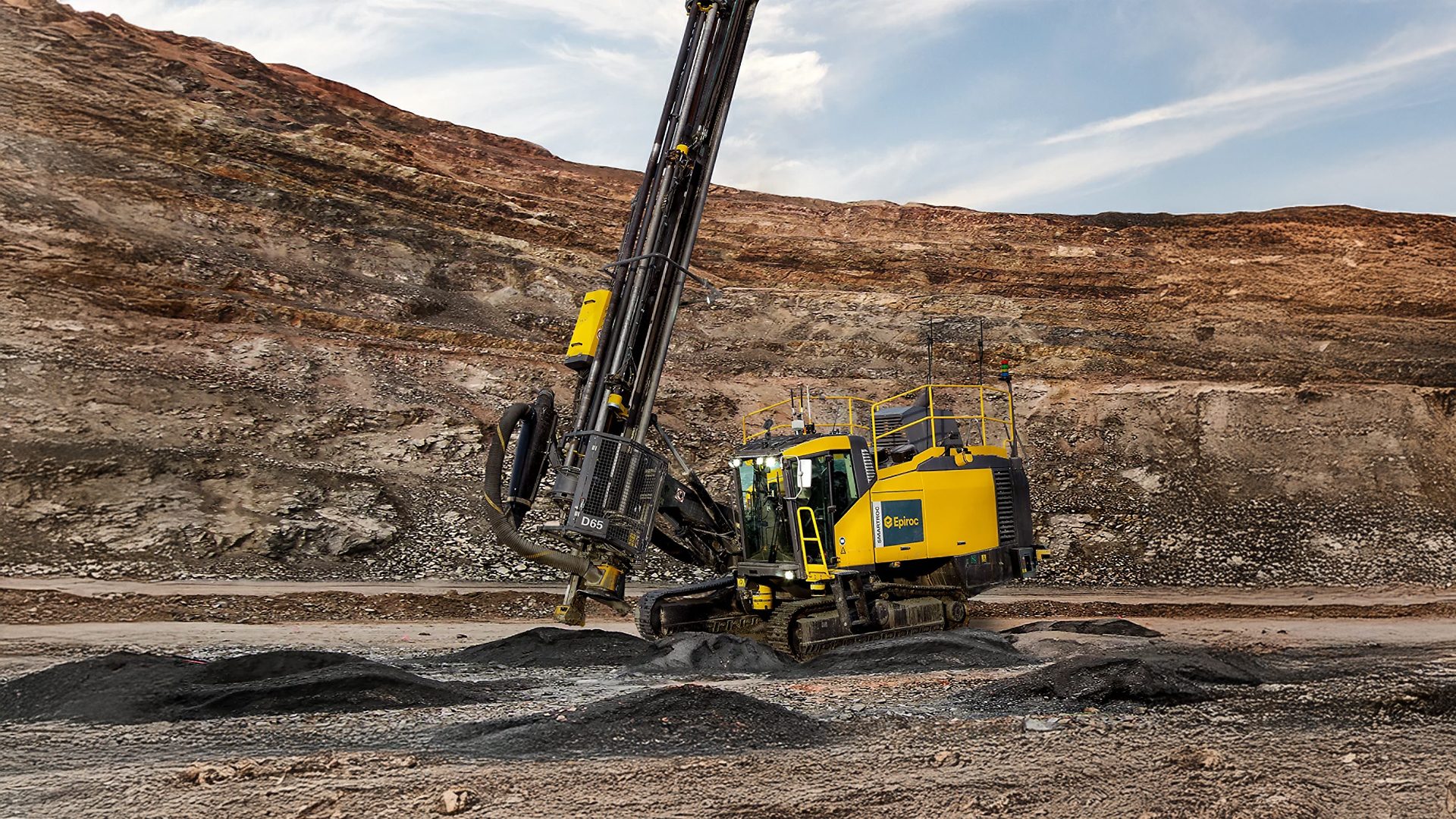 Yellow drilling rig working in an open-pit mine with a layered rock face.