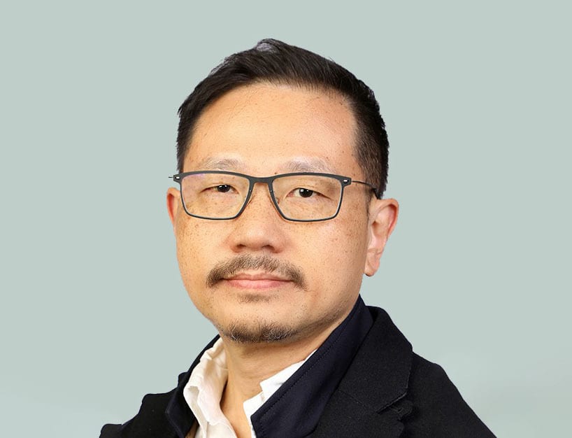 Headshot of an Asian man with glasses and a mustache, wearing a dark jacket over a white shirt, against a light background.
